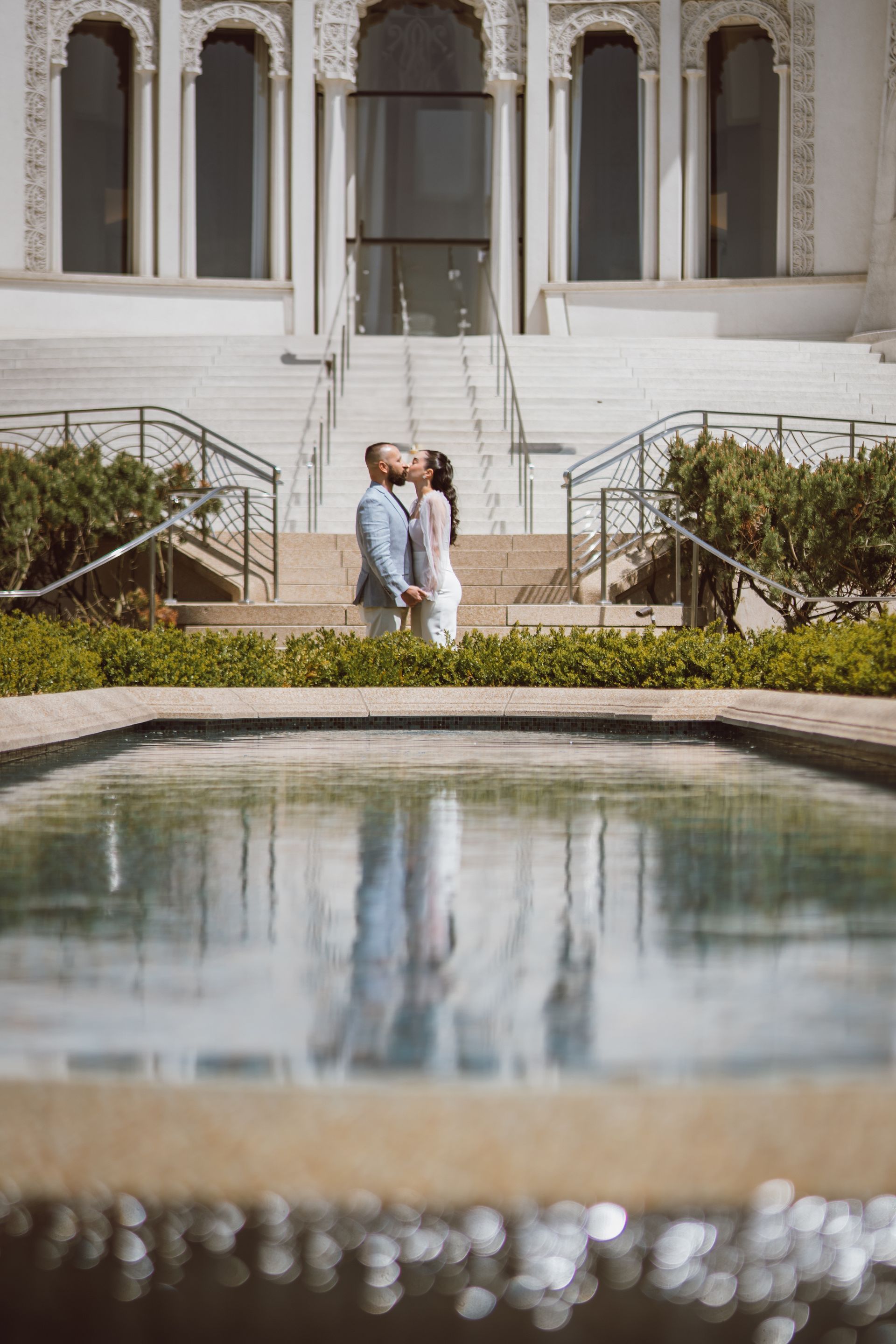 Couple kissing by a reflecting pool, in front of a white building with stairs.
