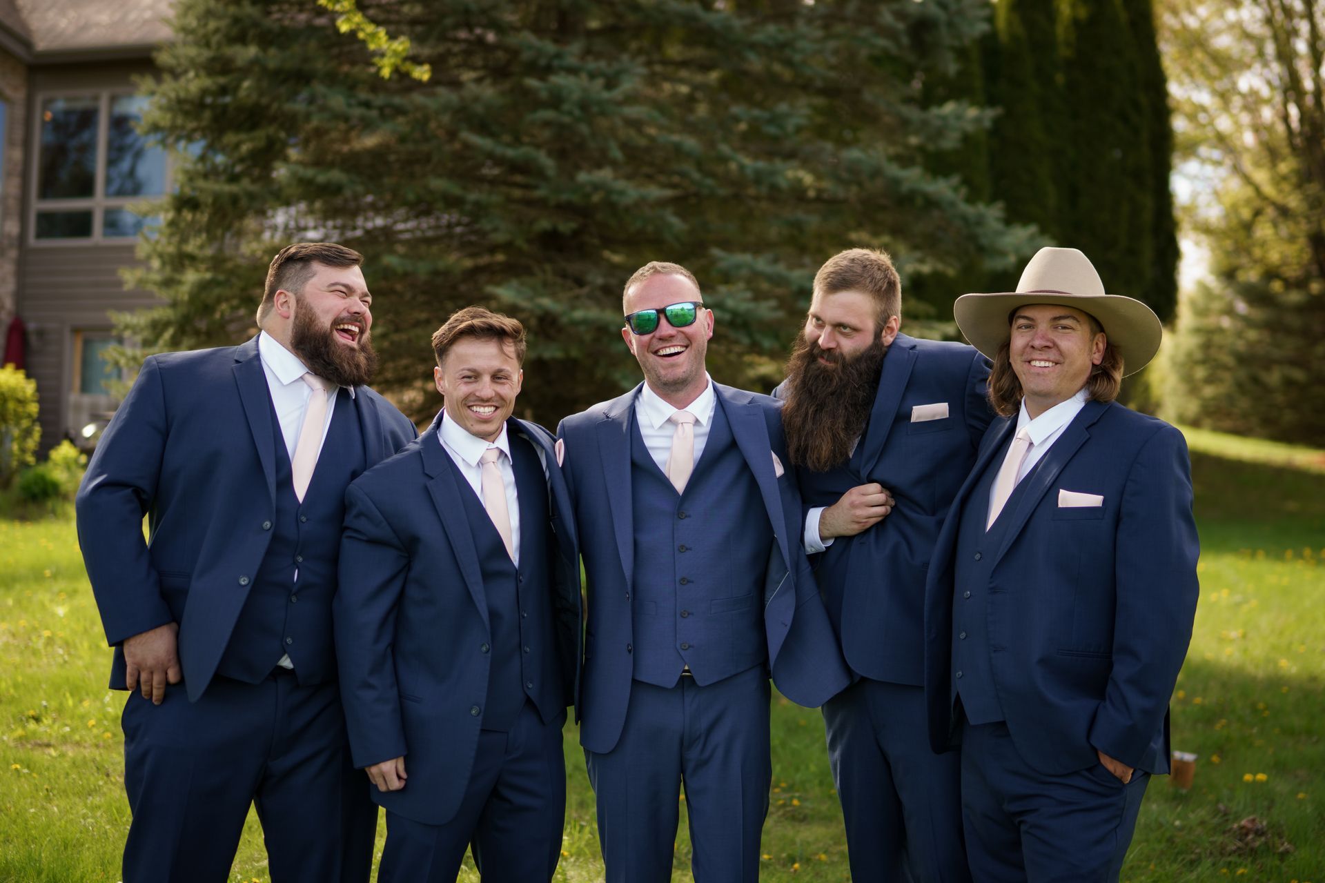 Men in navy suits outside; one wears a cowboy hat, some are laughing, sunny day.
