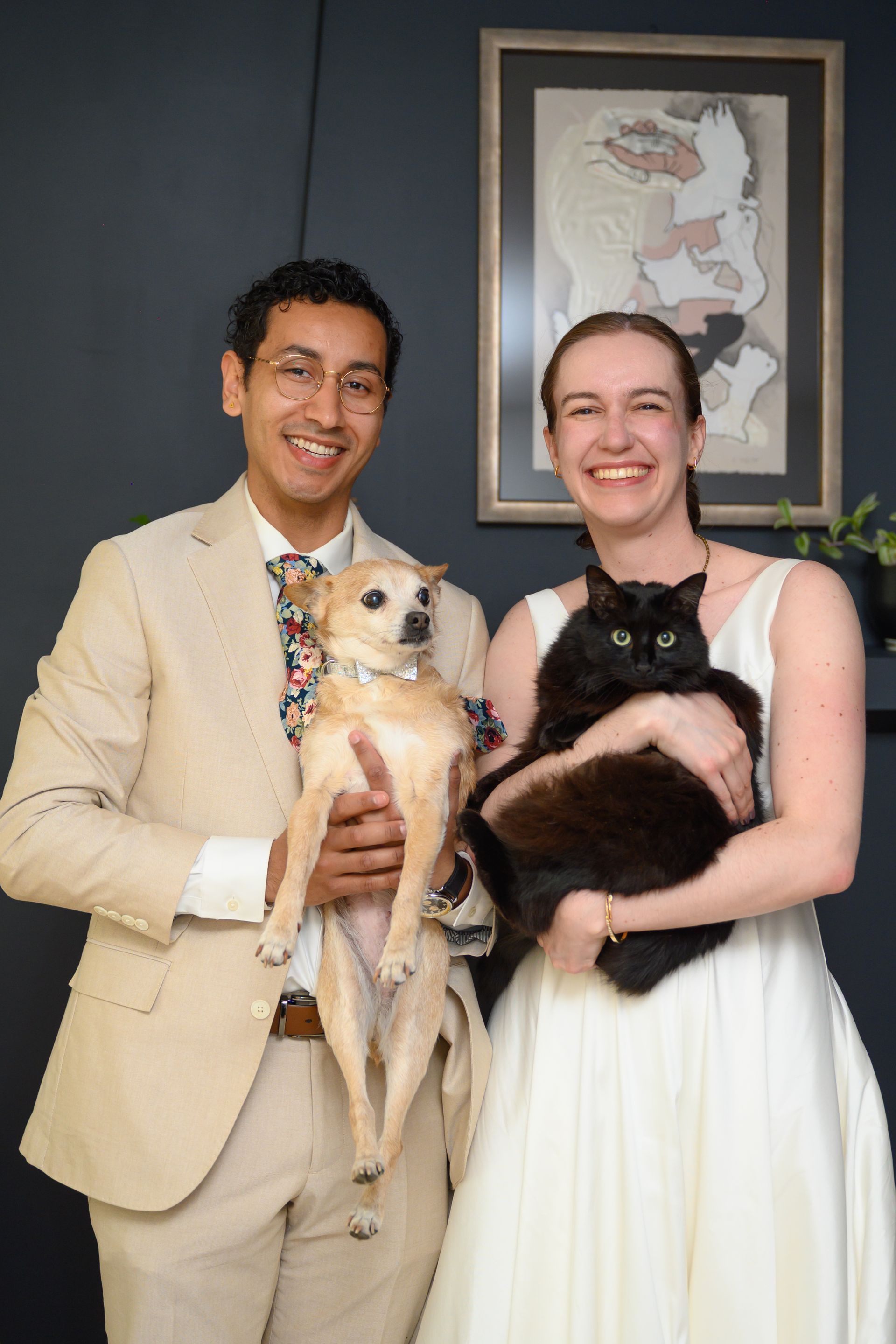Couple in wedding attire holding dogs, smiling. Neutral backdrop, art on wall.
