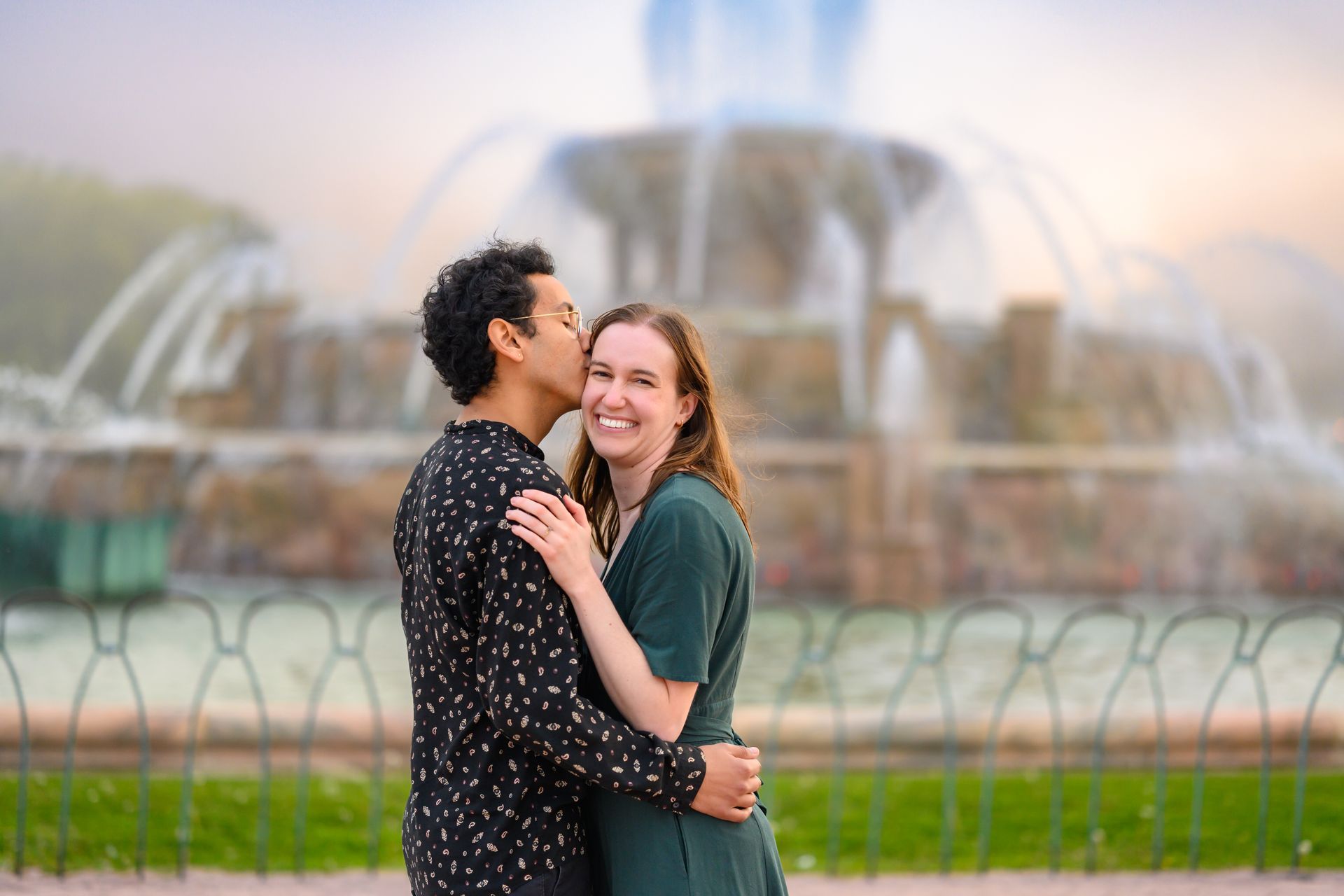 Couple hugging and kissing by a fountain; woman smiles, man kisses her cheek. Green dress, dark shirt.