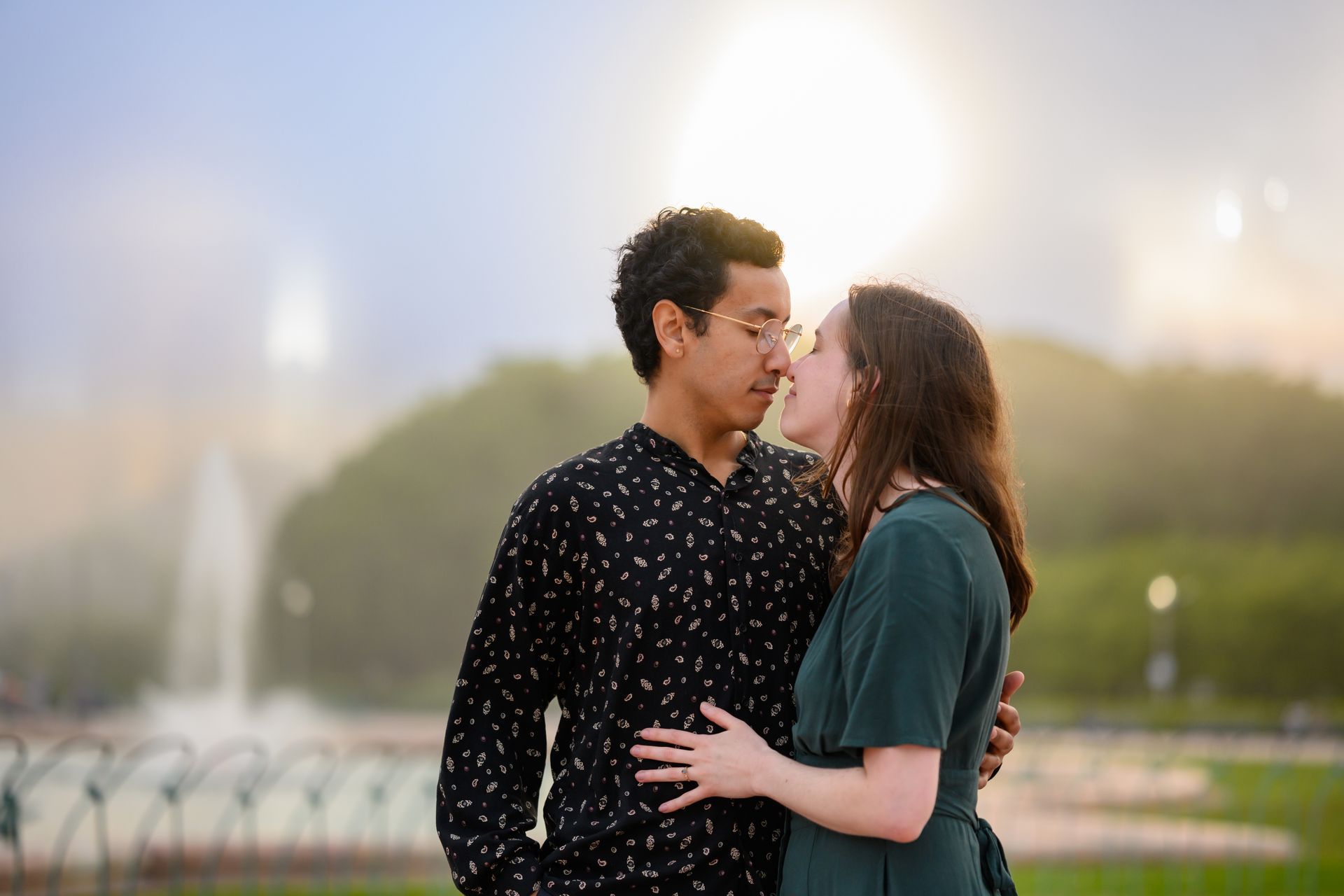Couple embracing outdoors, about to kiss. Fountain in background, soft light.