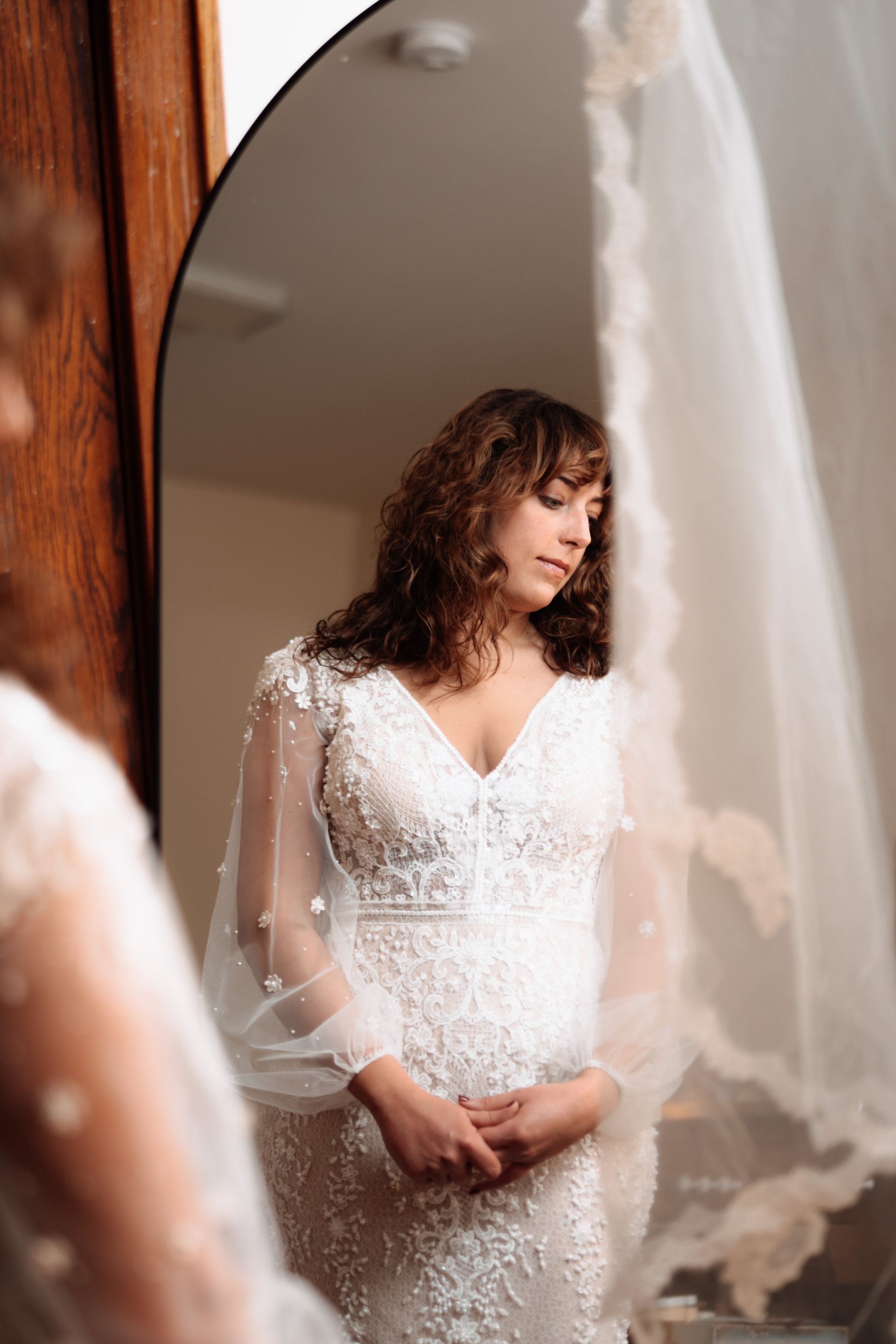 Bride in wedding dress, looking in mirror. She has curly hair and is wearing a long-sleeved gown, with a veil.
