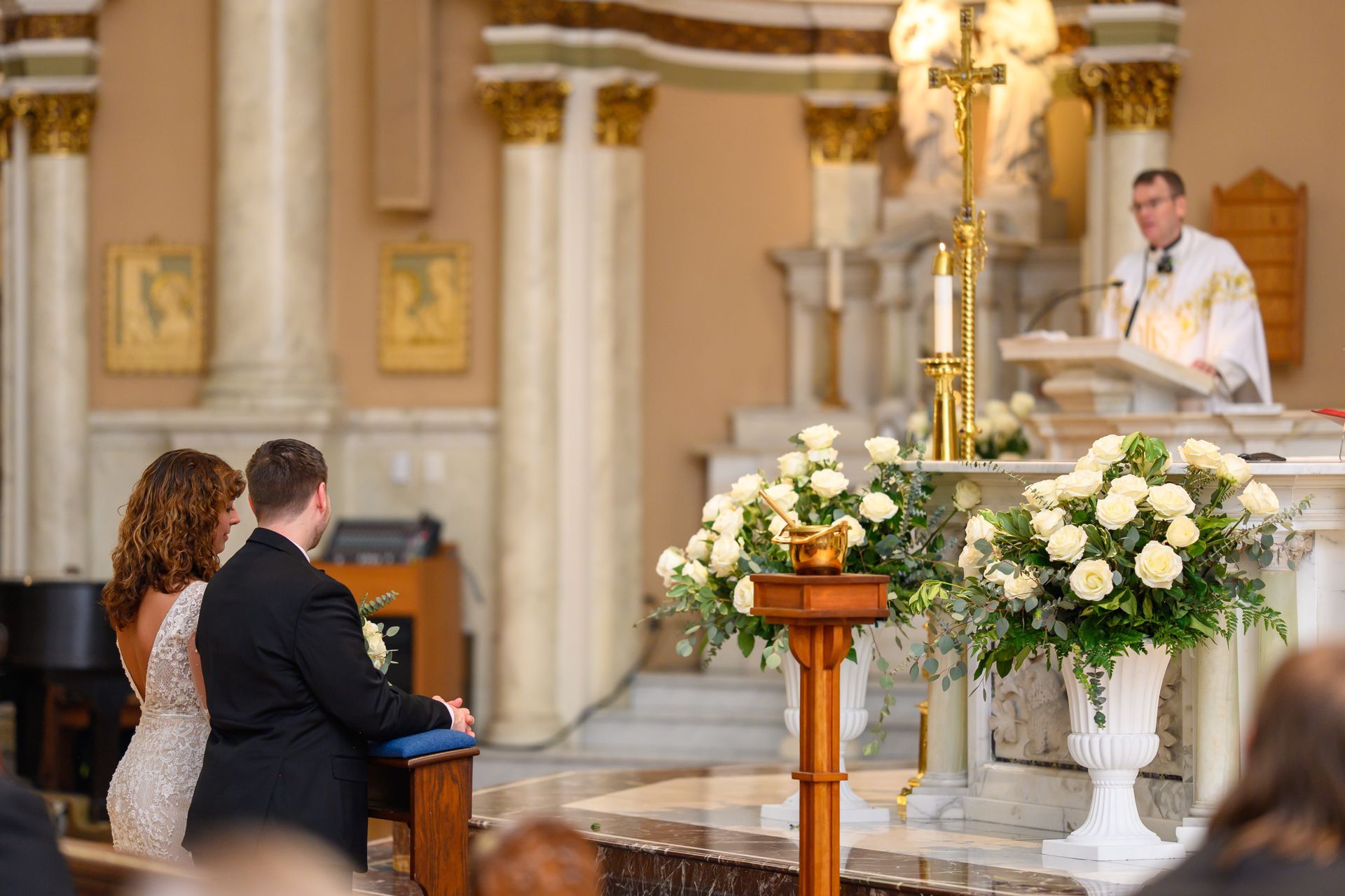 Couple at altar during a wedding ceremony with priest and floral arrangements.