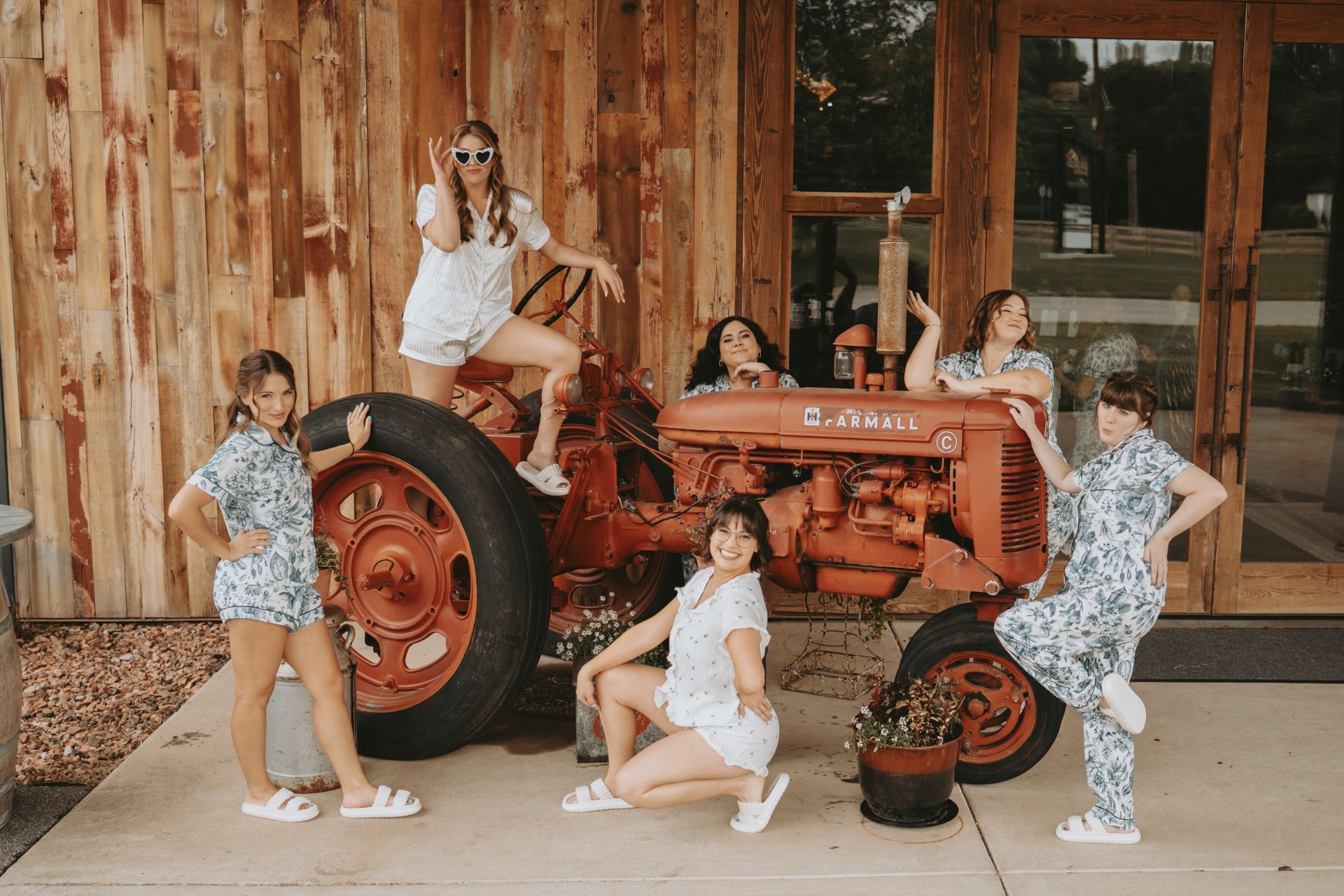 Six women in matching floral pajamas pose on and around a red tractor outside a wooden building.