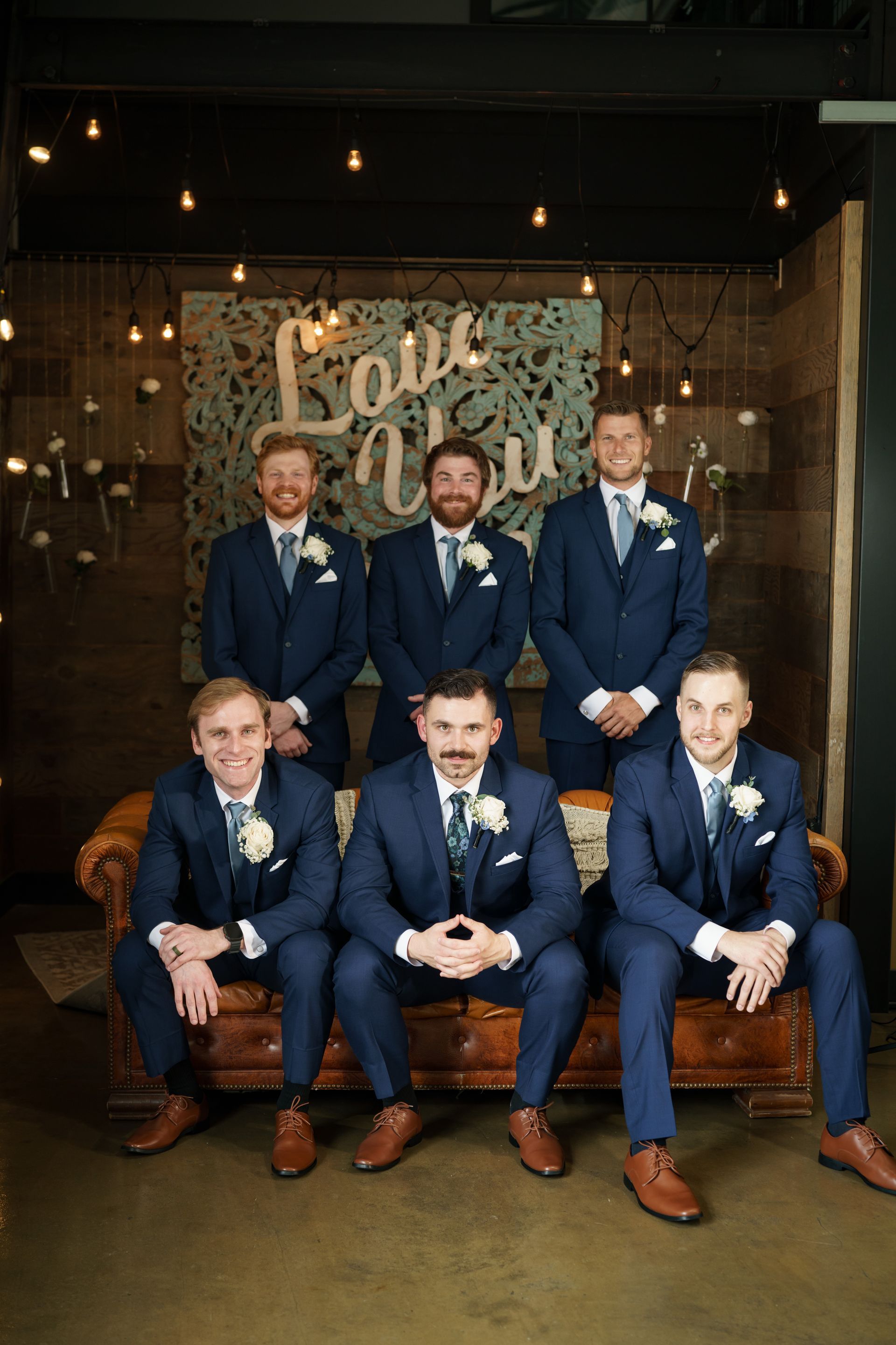 Groomsmen in navy suits pose for a photo. They stand and sit on a brown sofa, with 