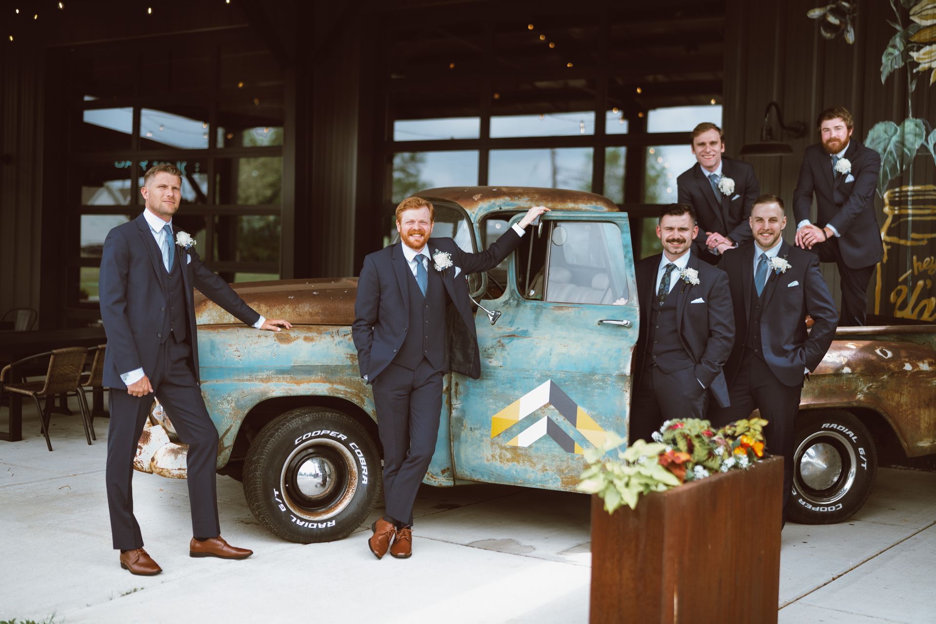 Groomsmen in navy suits pose near a vintage teal truck decorated with flowers. Building background.
