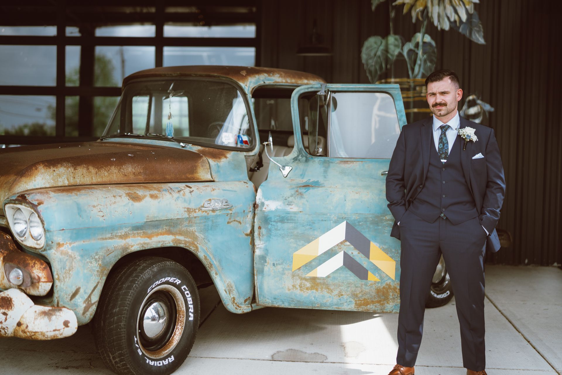 Man in a blue suit stands beside a weathered, light-blue pickup truck with the door open, in a modern setting.