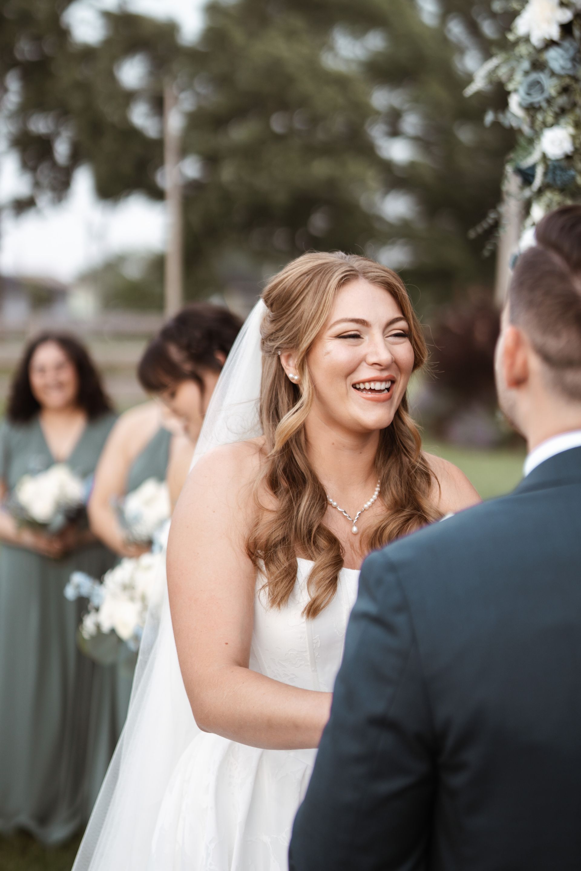 Bride smiles during outdoor wedding ceremony. Bridesmaids in green dresses, groom in blue suit.
