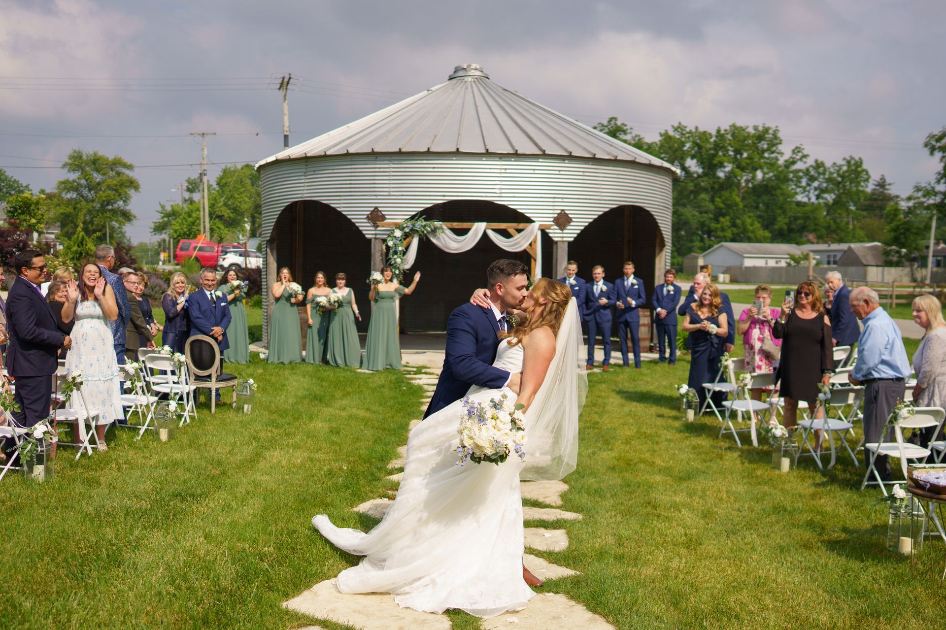 Newlyweds kissing at outdoor ceremony, guests watch. Silo structure in background, green grass, sunny day.