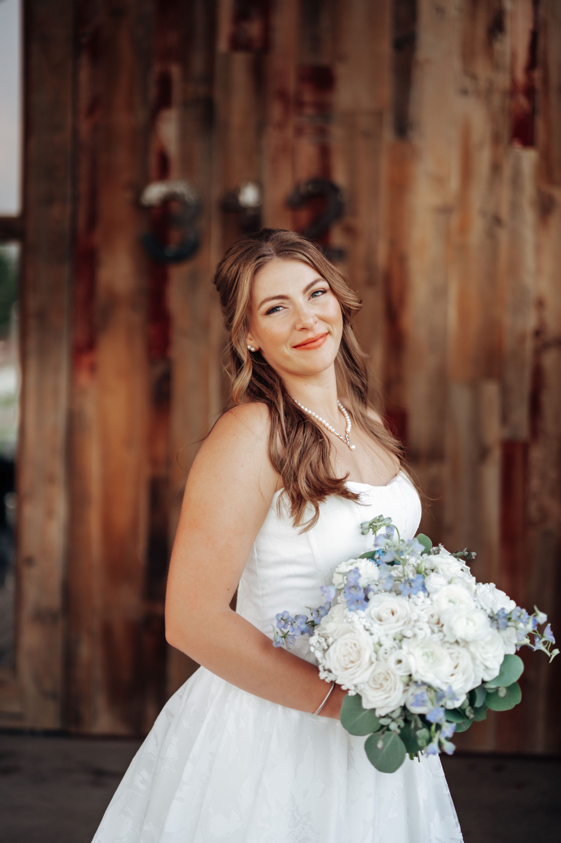 Bride in white dress holding bouquet, smiling; wood backdrop with wreaths.