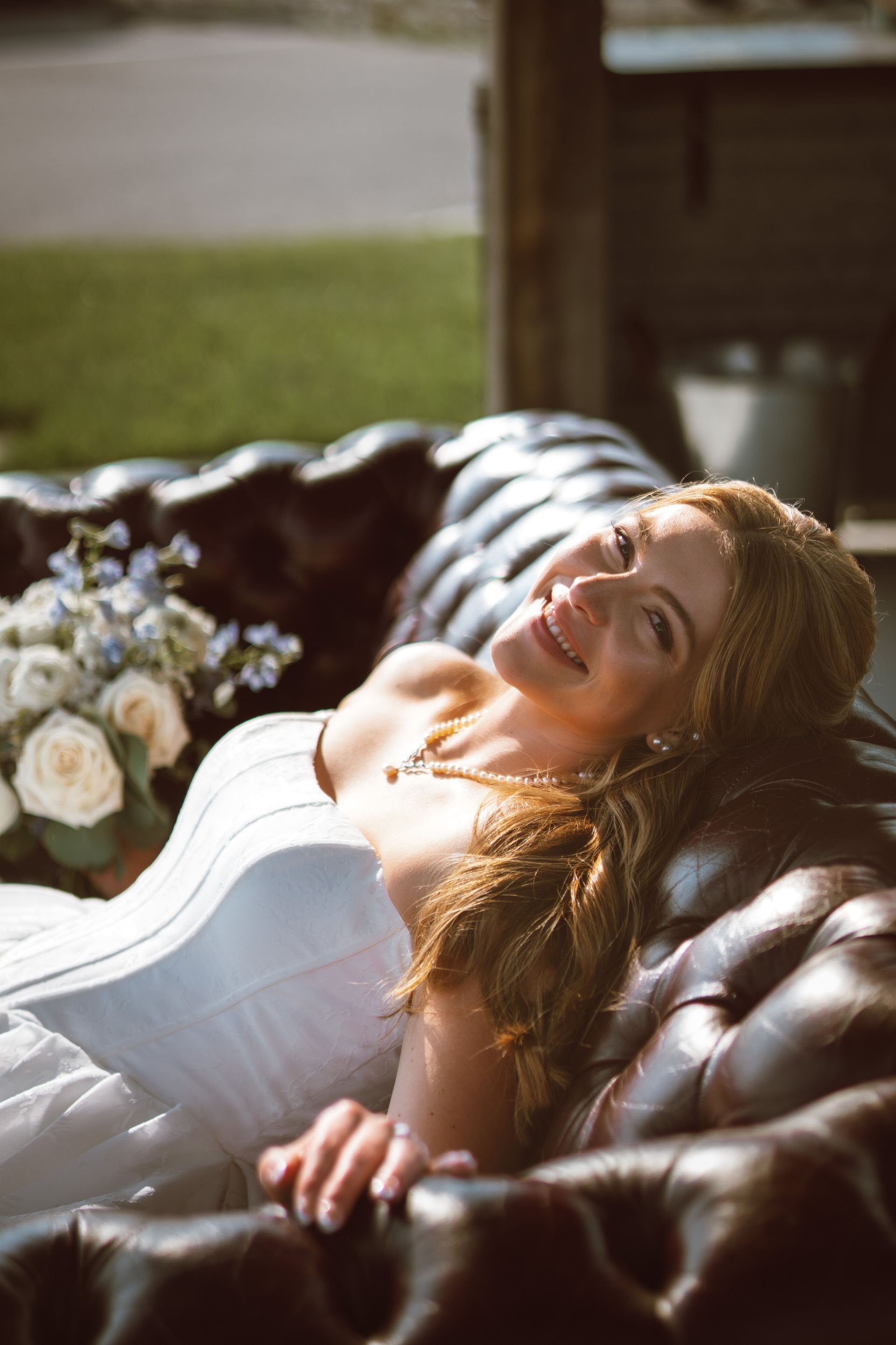 Bride in white gown reclines on a brown tufted leather couch, smiling with bouquet.