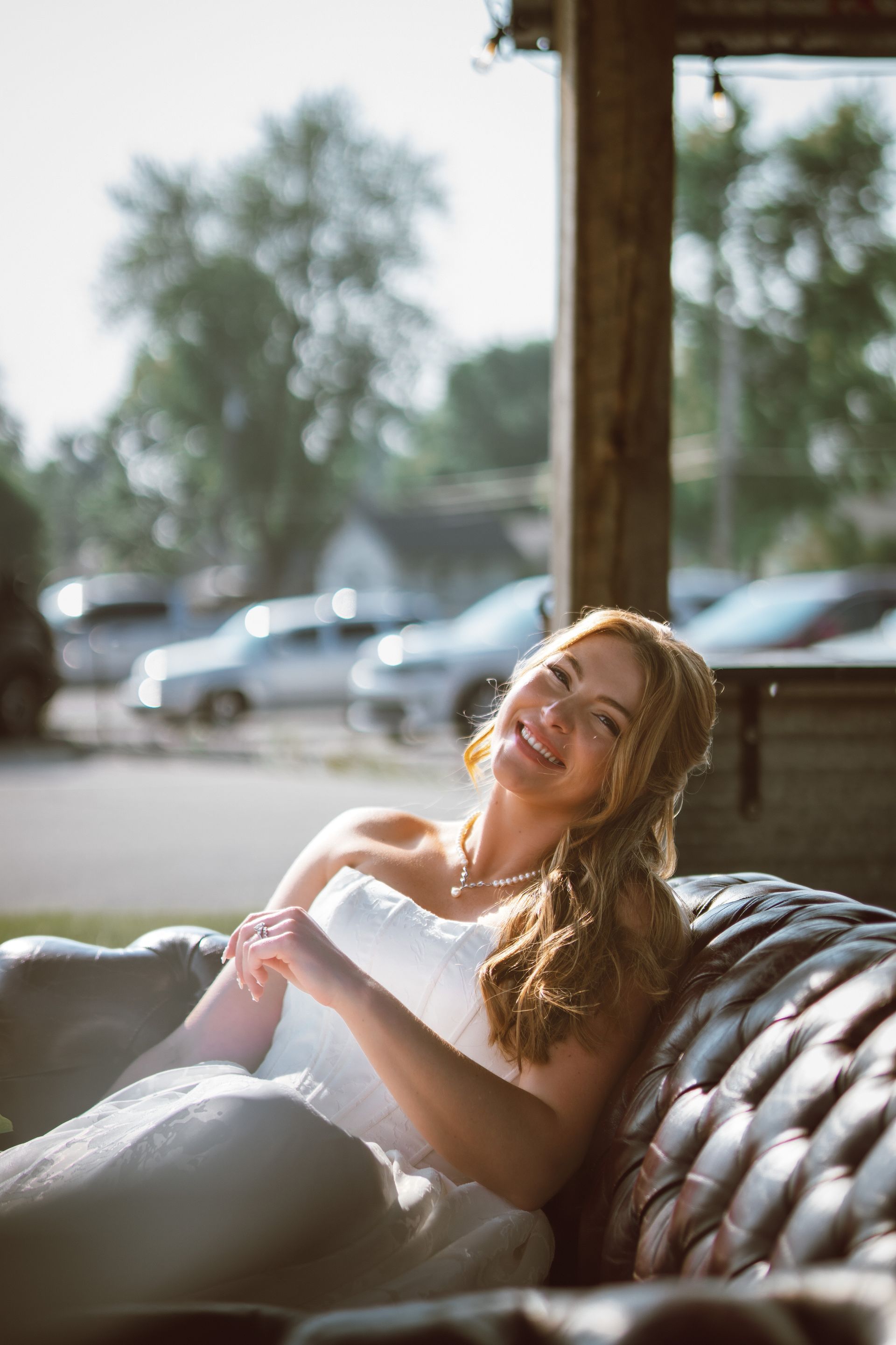 Bride in strapless white dress smiling, sitting on a tufted leather couch, bright sunlit outdoors.