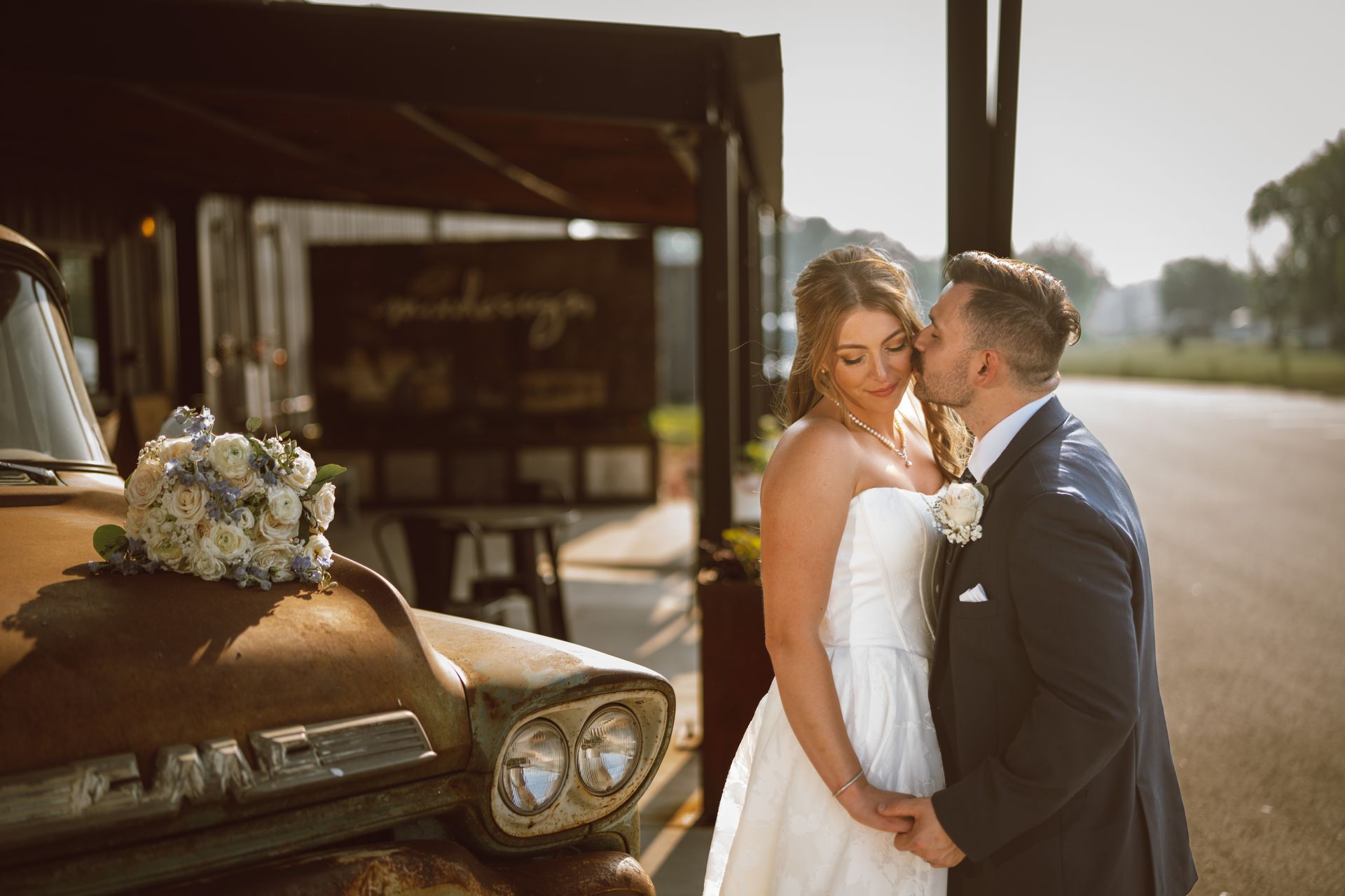 Bride and groom embrace by a rusty truck. The groom kisses the bride's cheek. Floral arrangement on the hood.