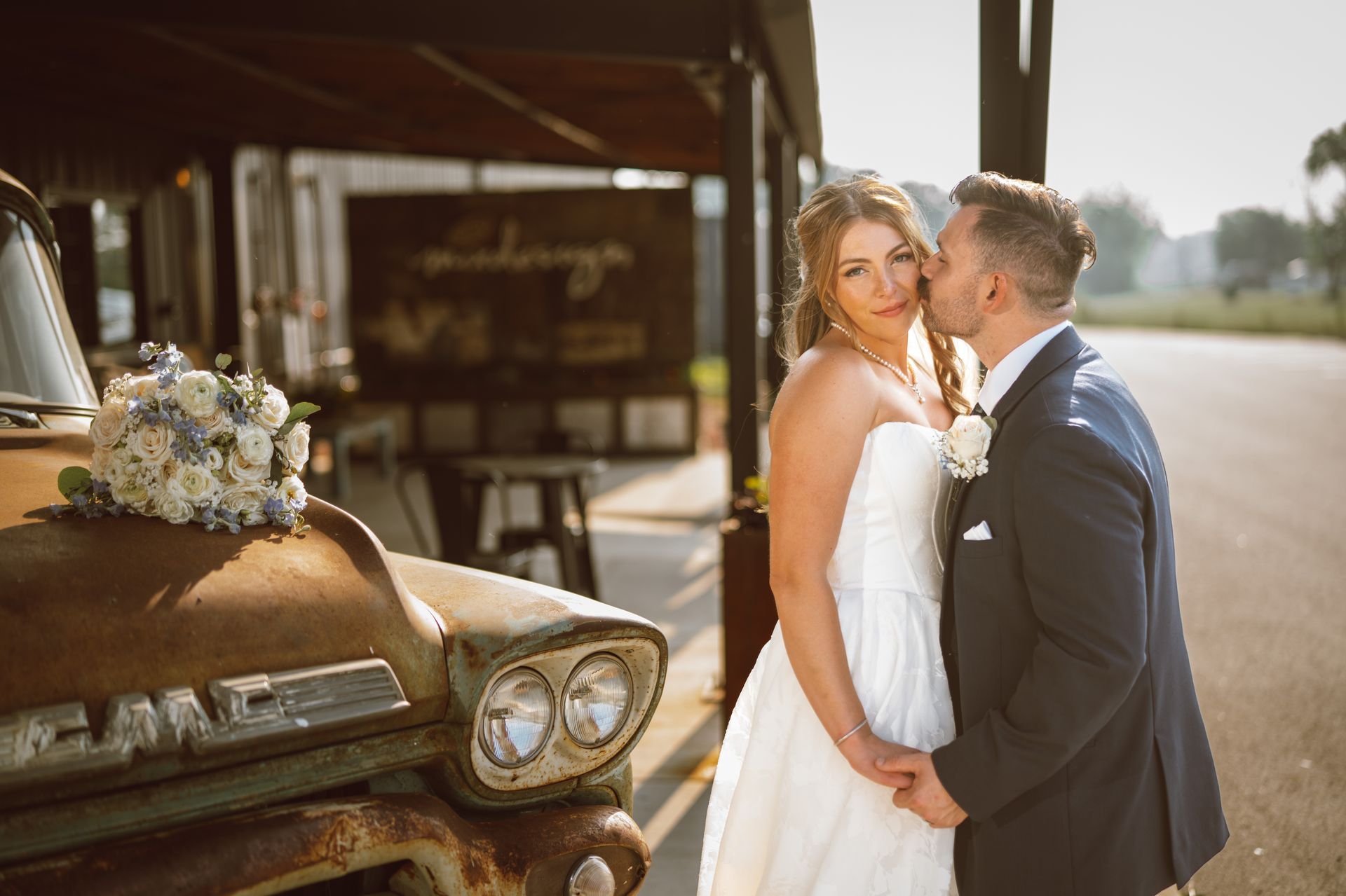 Bride and groom pose by a rusty truck, groom kissing the bride's cheek. Wedding setting, sunny day.