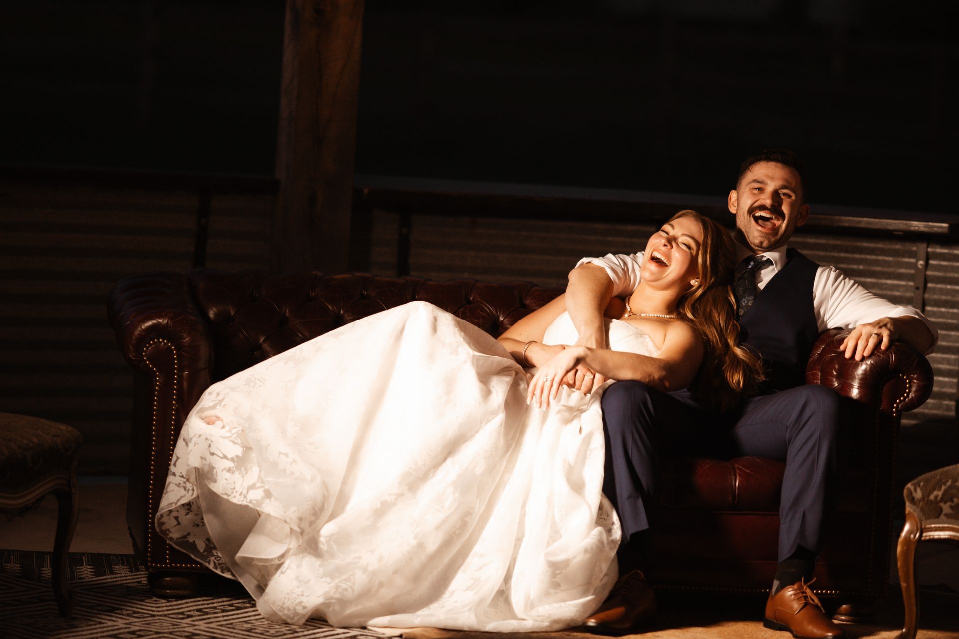 Bride and groom laughing on a leather couch; bride in white gown, groom in vest and tie. Dark setting.