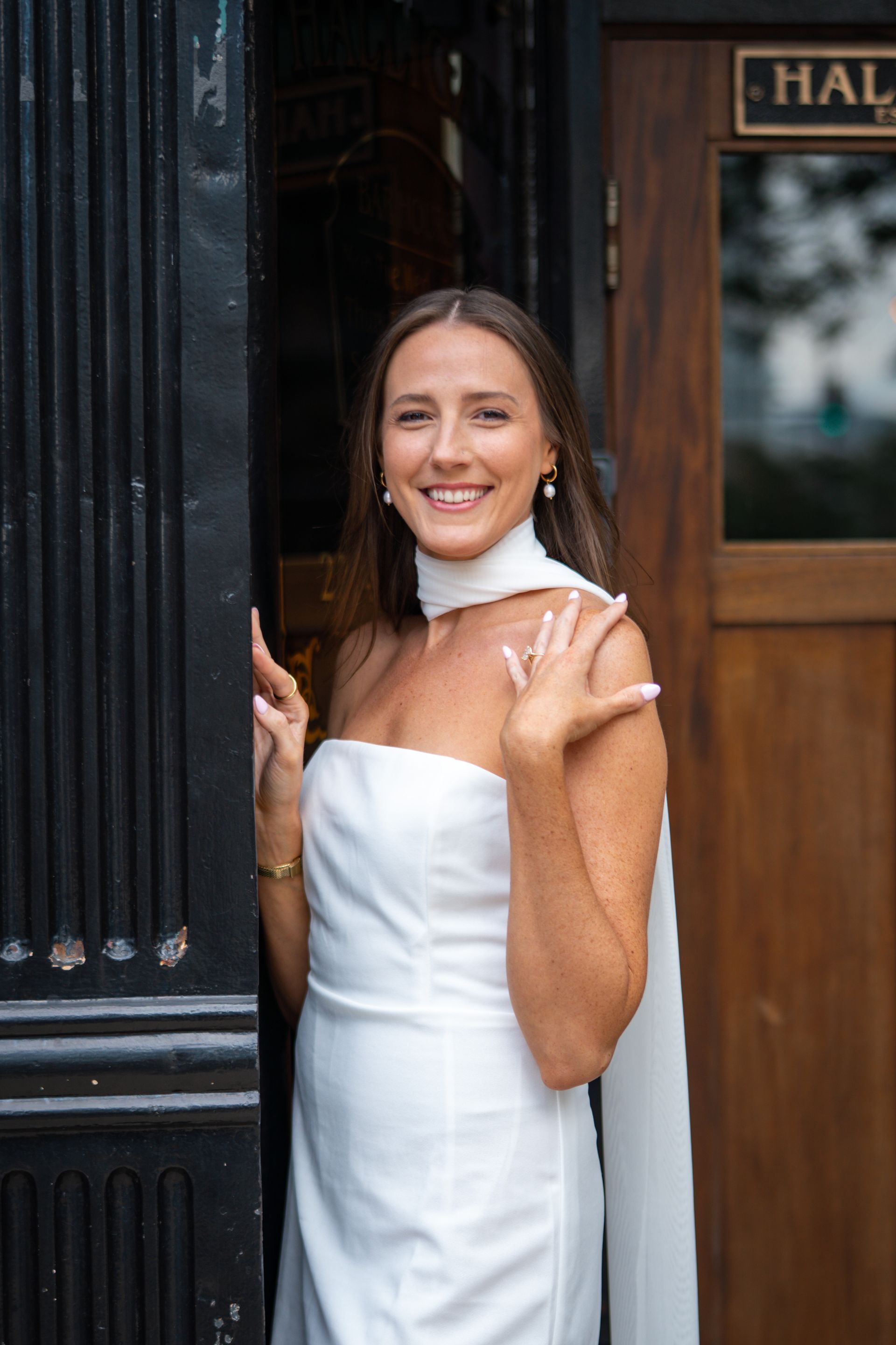 Woman in white dress smiles, standing in doorway, hand near face, near a door with a sign.