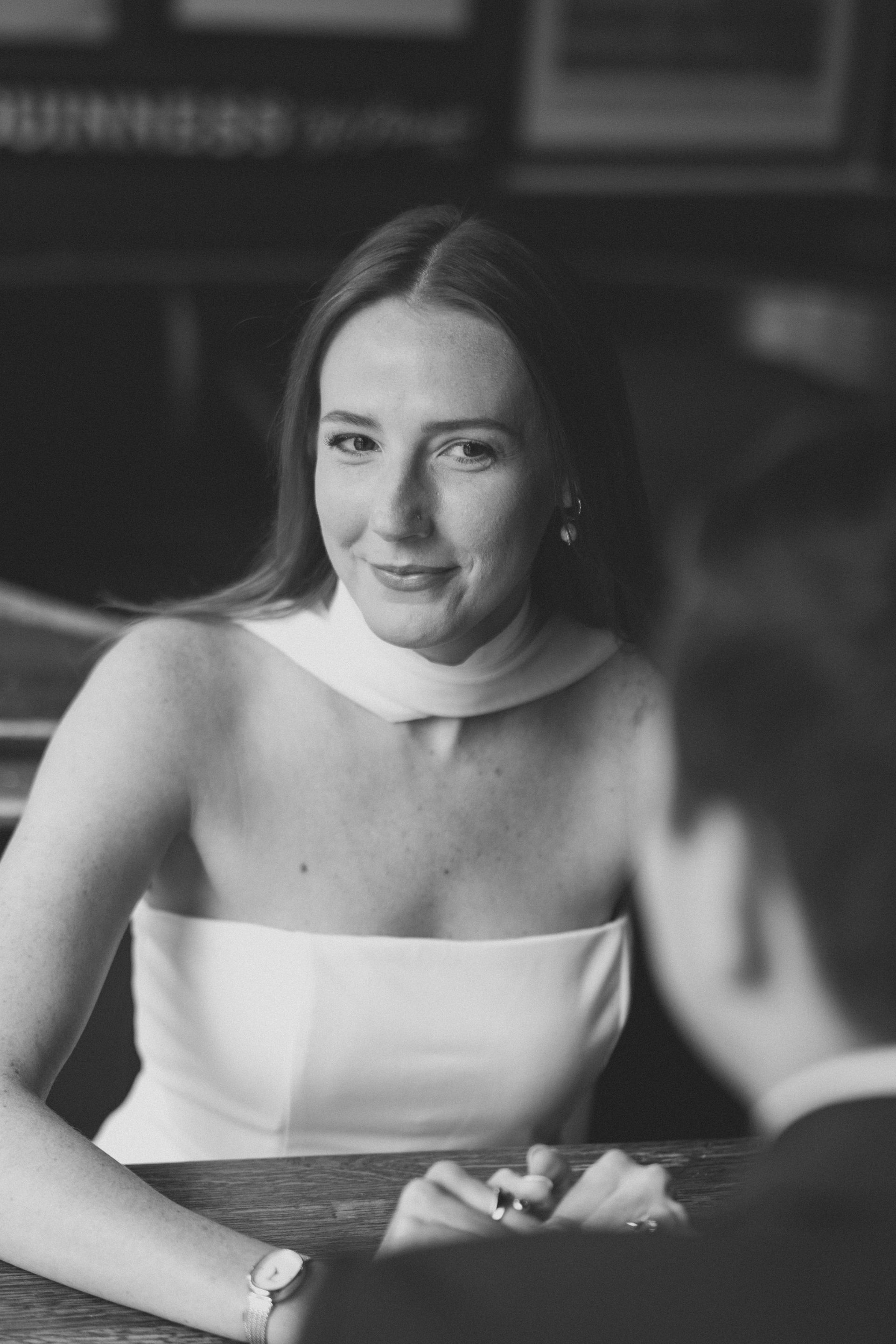 Woman in white dress smiles at person out of frame. Indoors, seated.