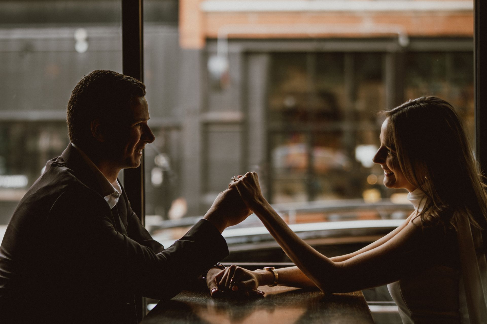 Couple holding hands, silhouetted at a table, looking at each other through a window.