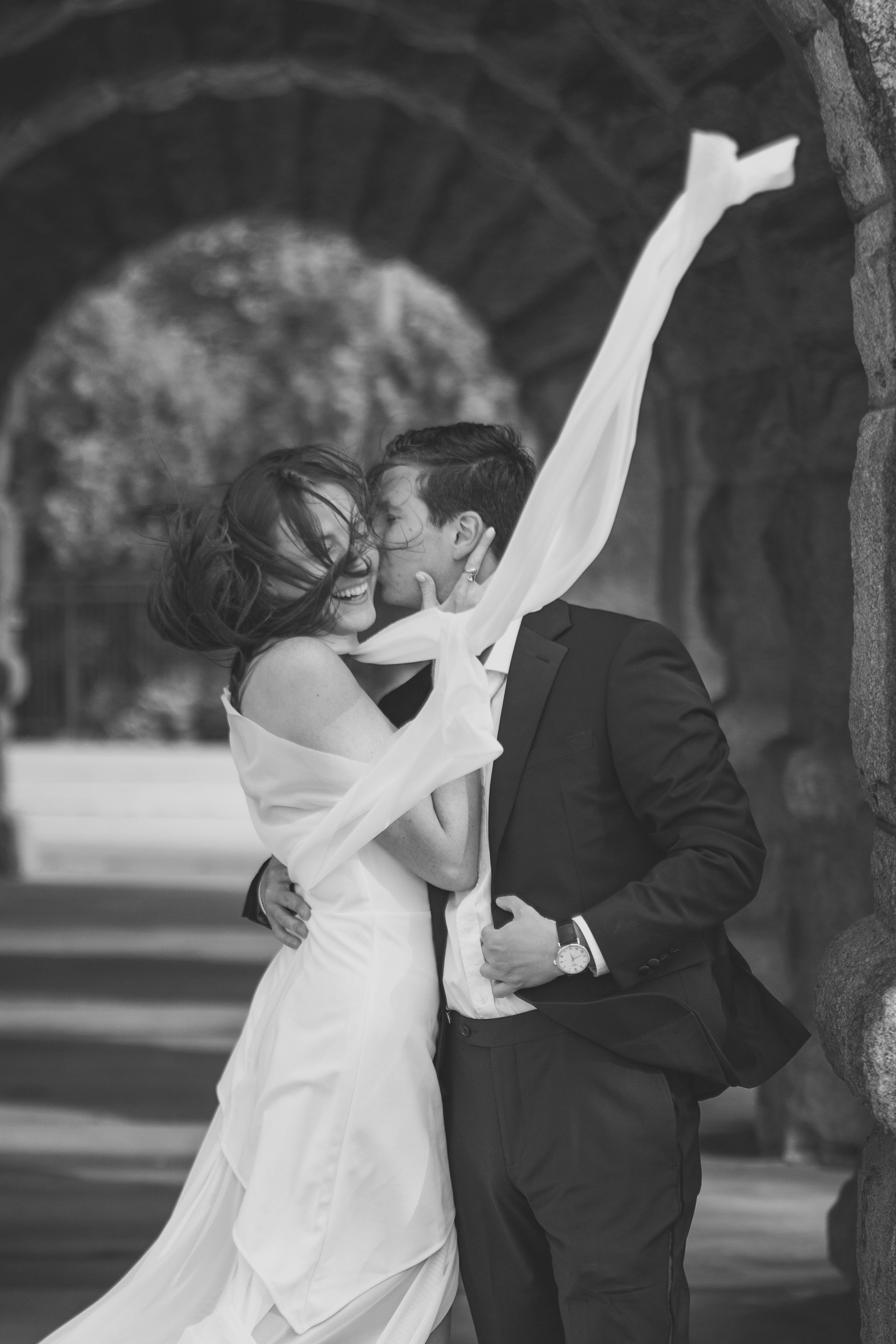 Couple embracing, with bride's scarf blowing in the wind, under a stone archway.