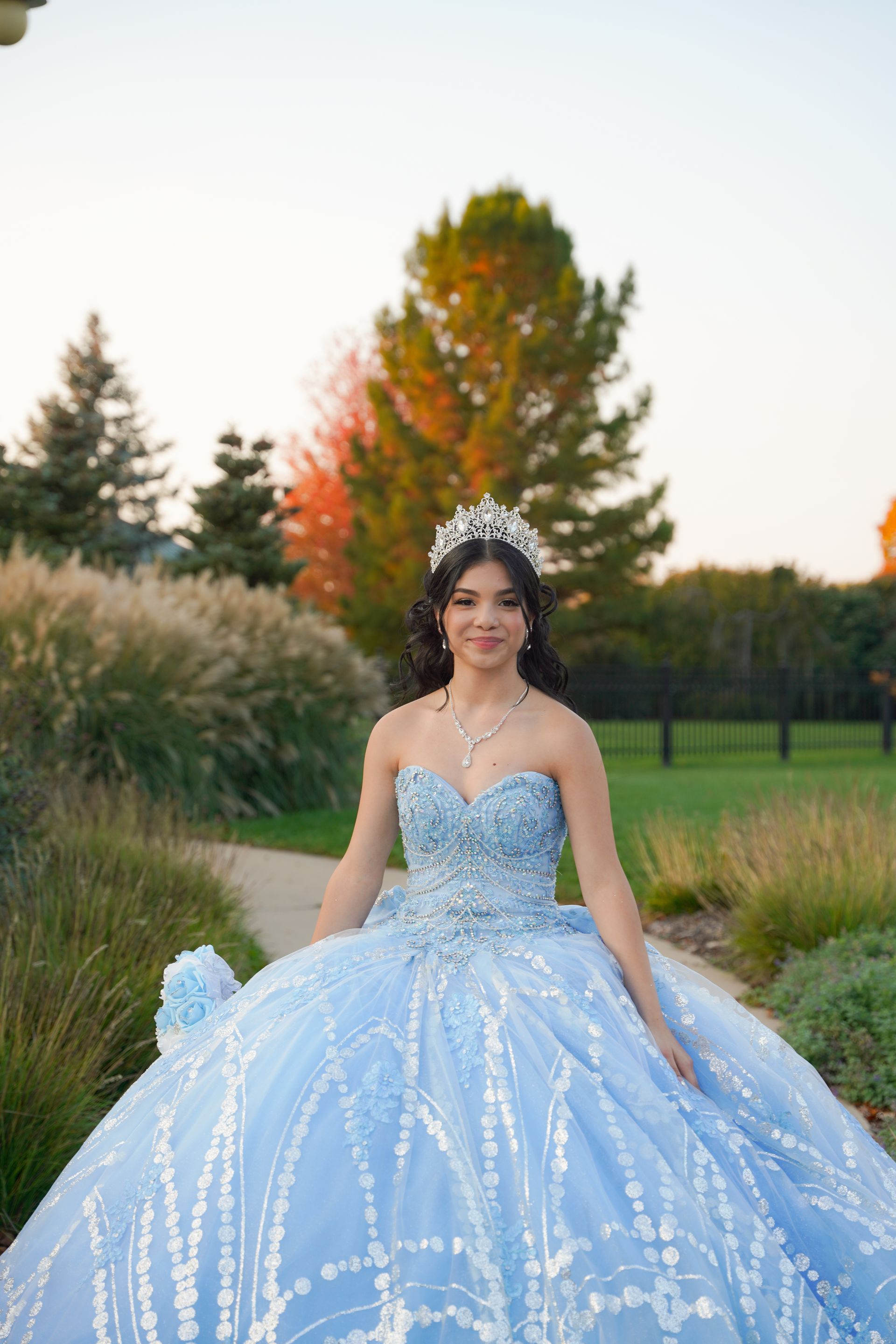 Woman in a blue formal gown, wearing a tiara, smiles outdoors.