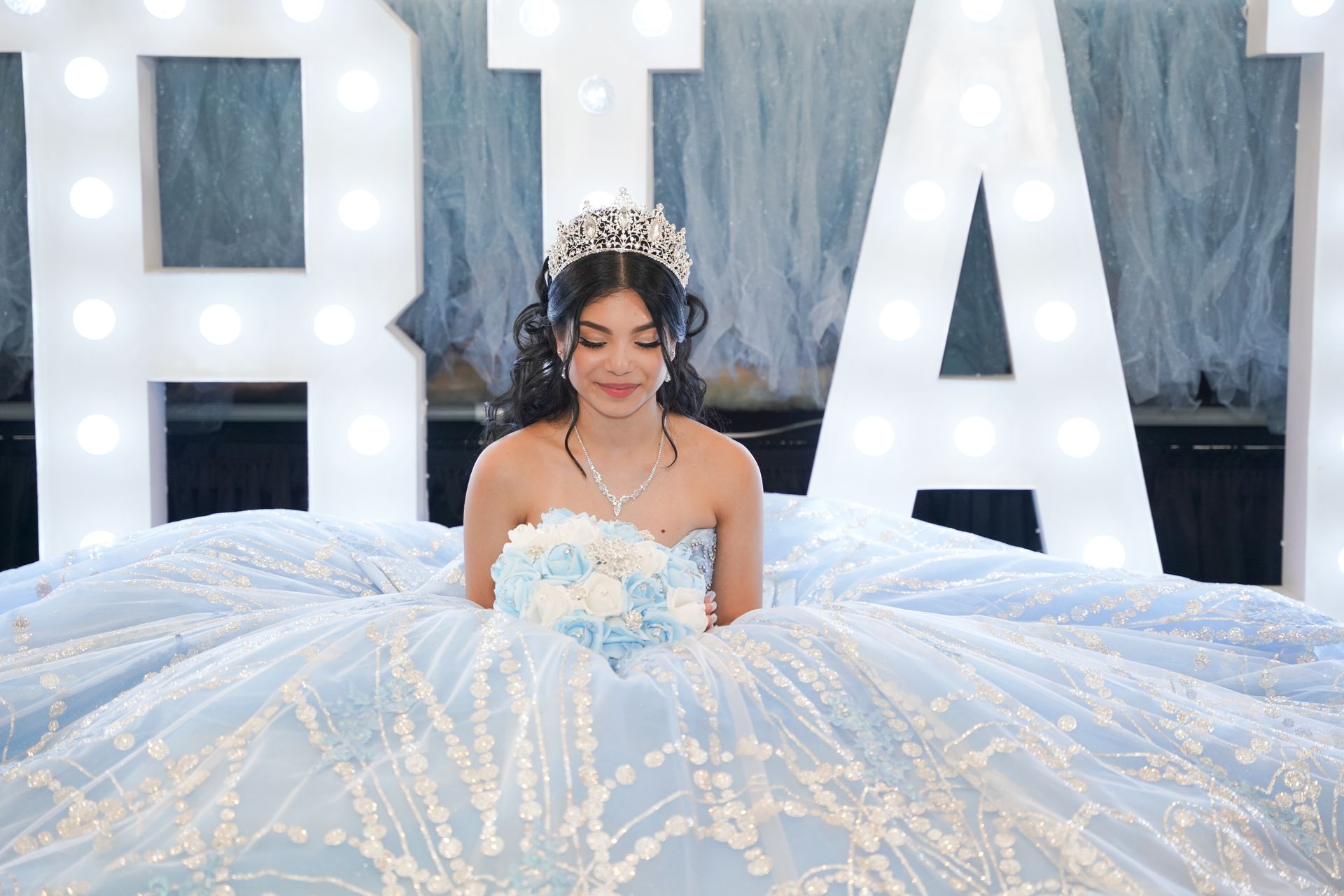 Girl wearing a crown and light blue ballgown, holding flowers. She's in front of illuminated letters spelling 