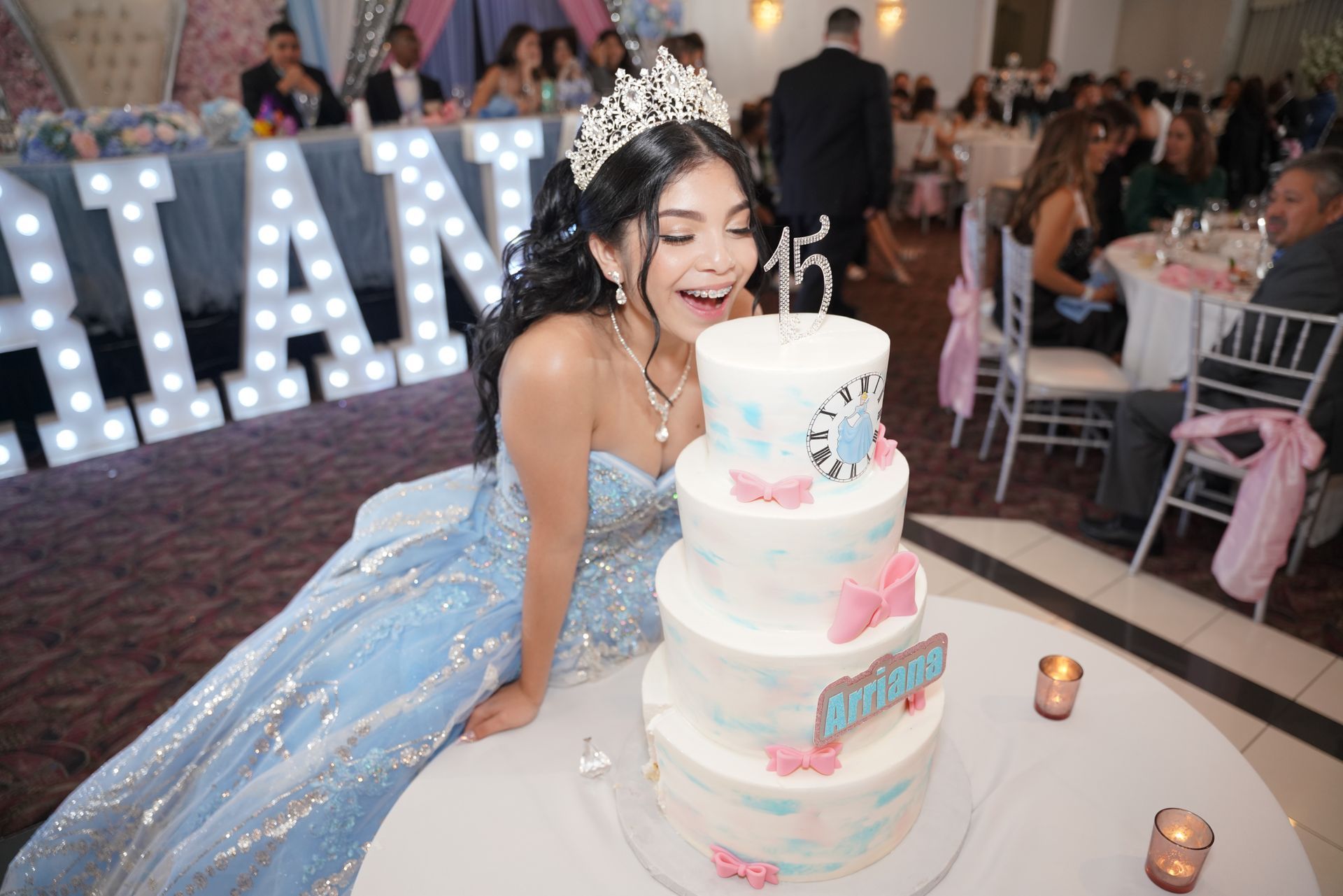Girl in a blue gown with a tiara blowing out candles on a four-tier cake at a party, 
