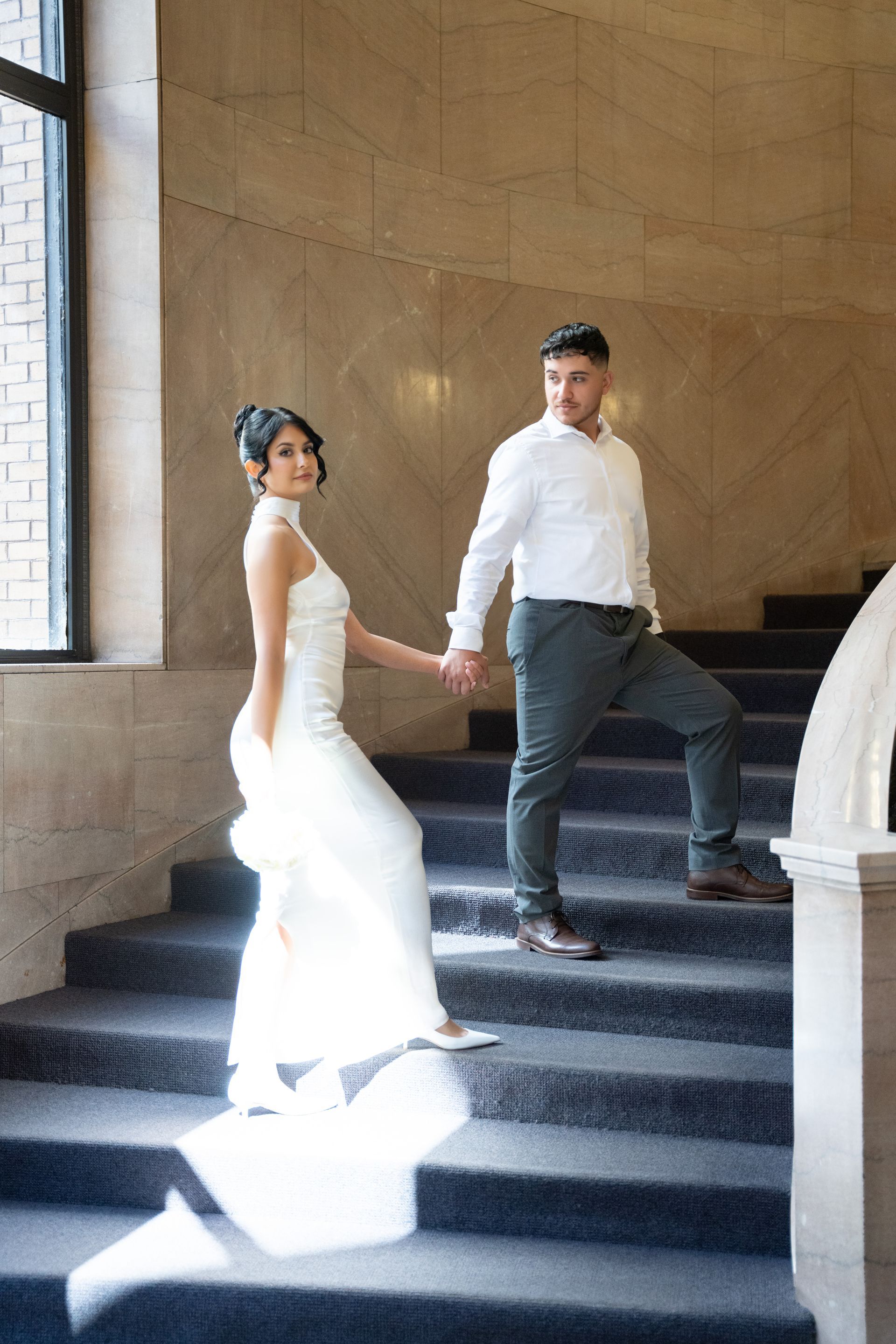Bride and groom holding hands, walking down marble staircase. Bride in white dress, groom in shirt and pants.