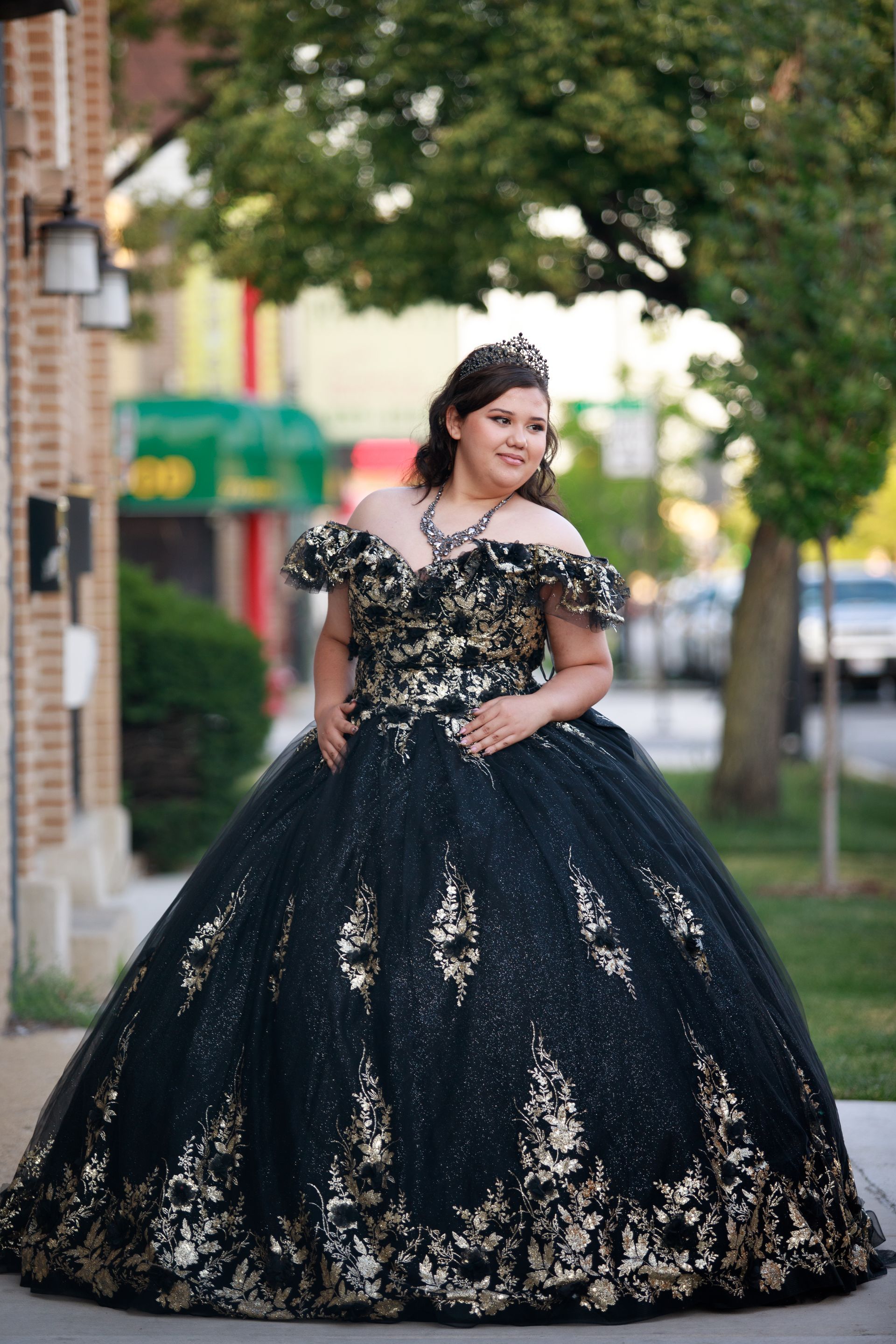 Woman in a black and gold ballgown with tiara, smiling, standing outdoors.