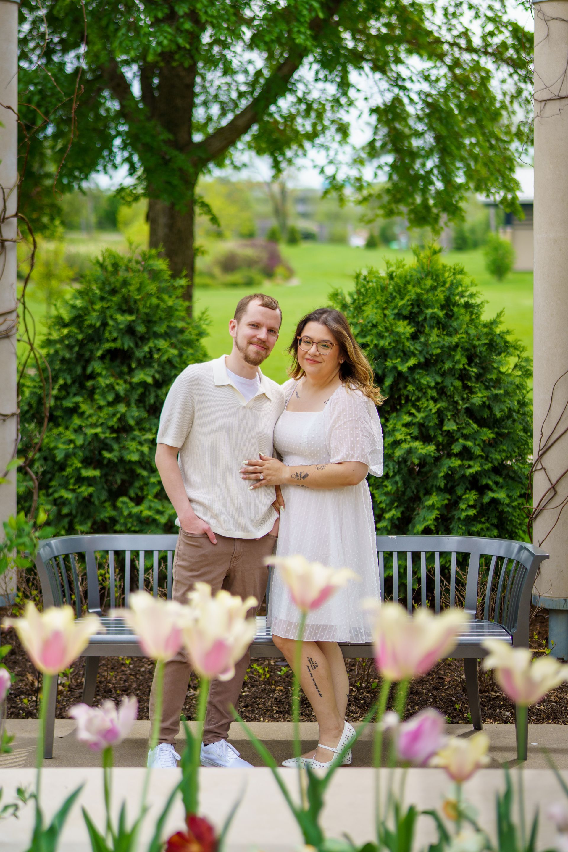 Couple standing in garden, smiling. Woman in white dress, man in tan shirt. Tulips in foreground.