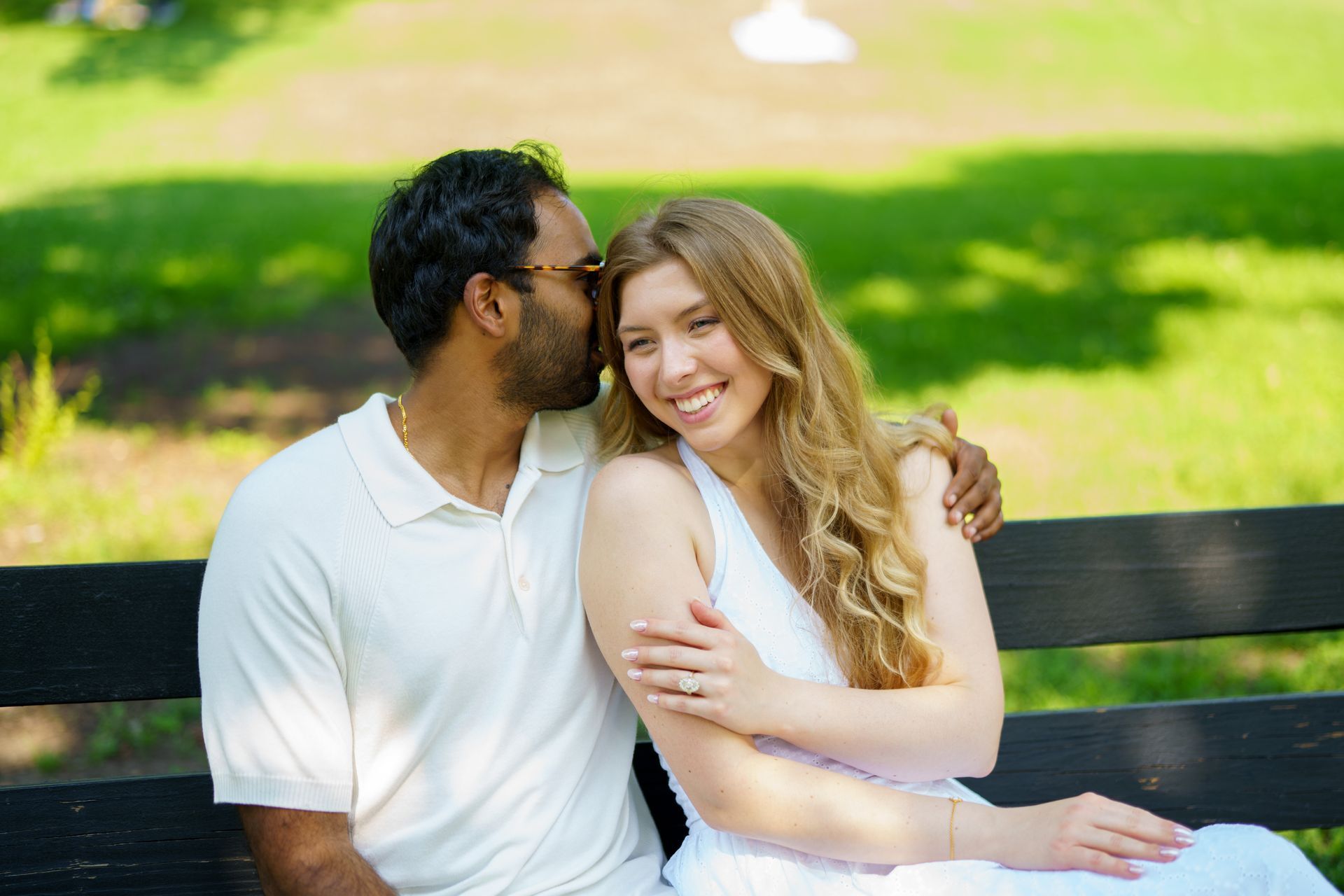 Couple sitting on a park bench. Man kisses woman's cheek. She smiles. Green trees in the background.