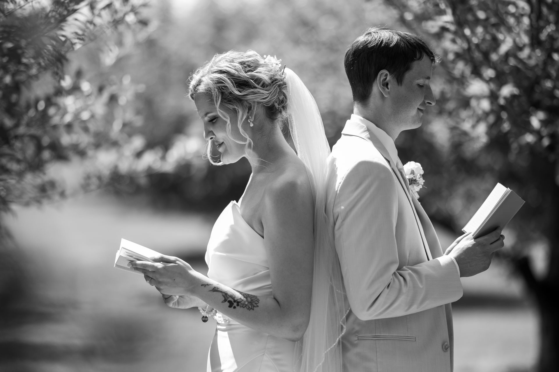 Wedding couple, in formal attire, back-to-back, reading from books outdoors.