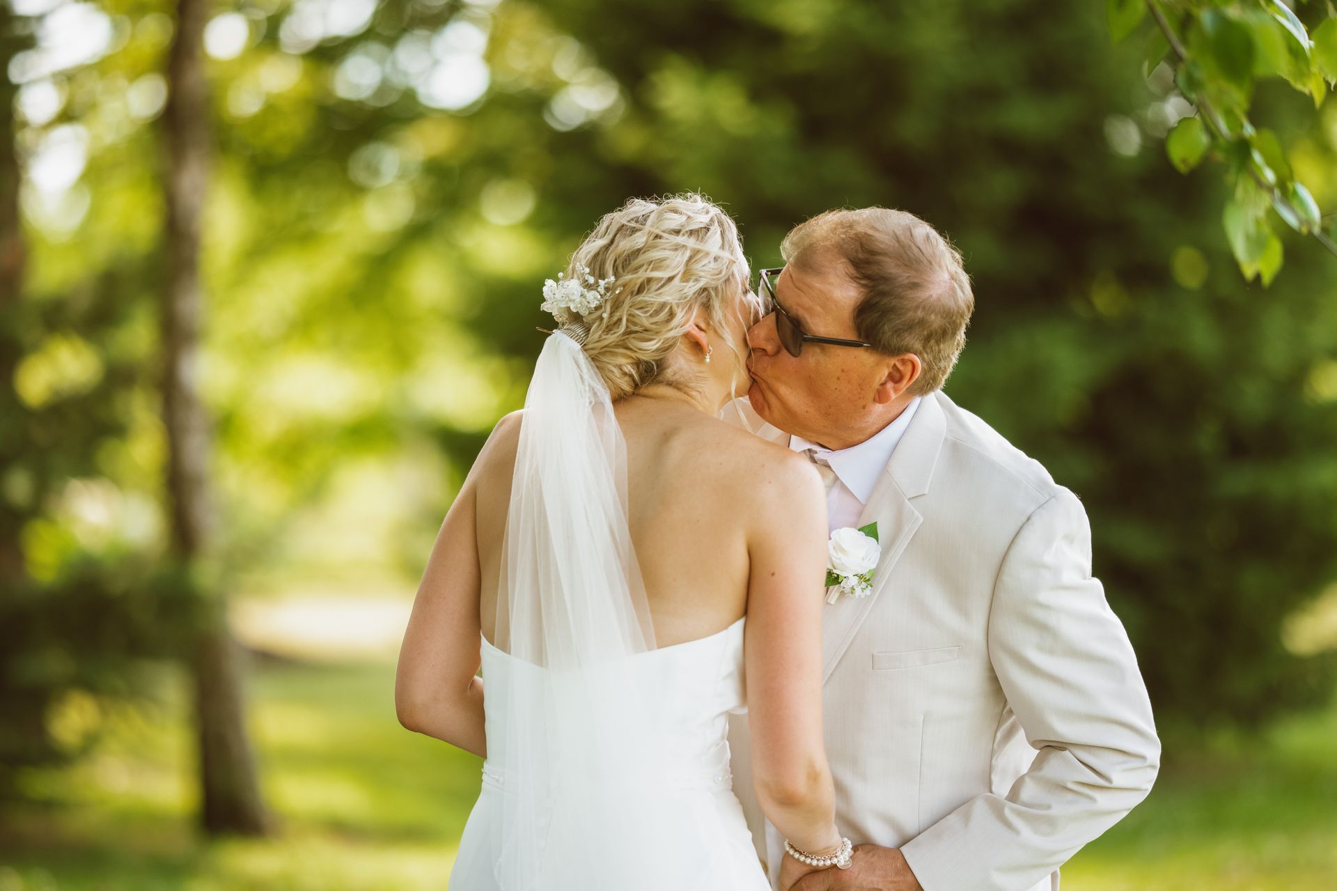 Bride kisses a man's cheek, outdoors. The bride is in a white dress and veil; the man in a tan suit.