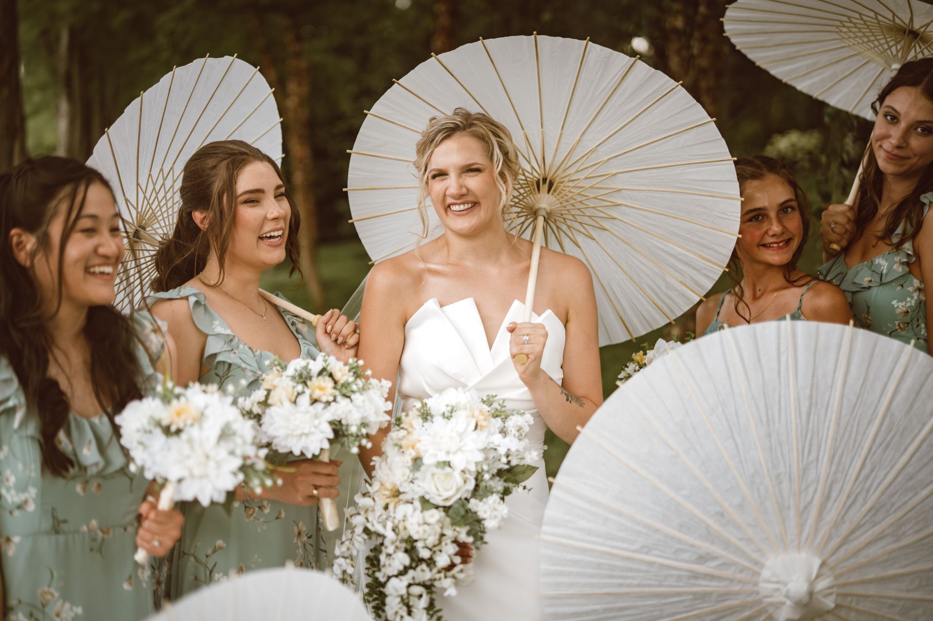 Bride and bridesmaids smiling with parasols and bouquets in a park setting.