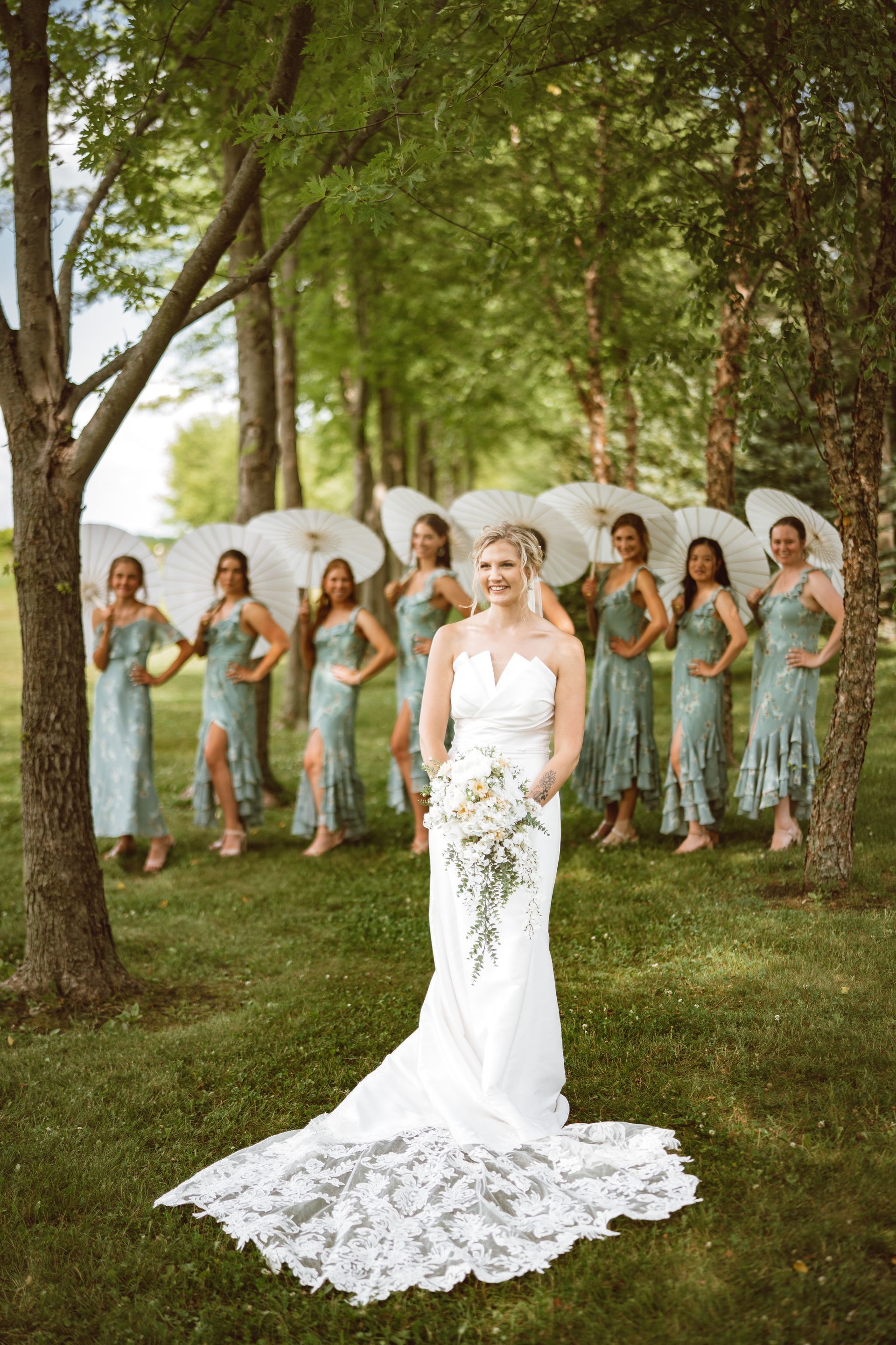 Bride in white gown with bouquet, and bridesmaids in green dresses, holding parasols outdoors.