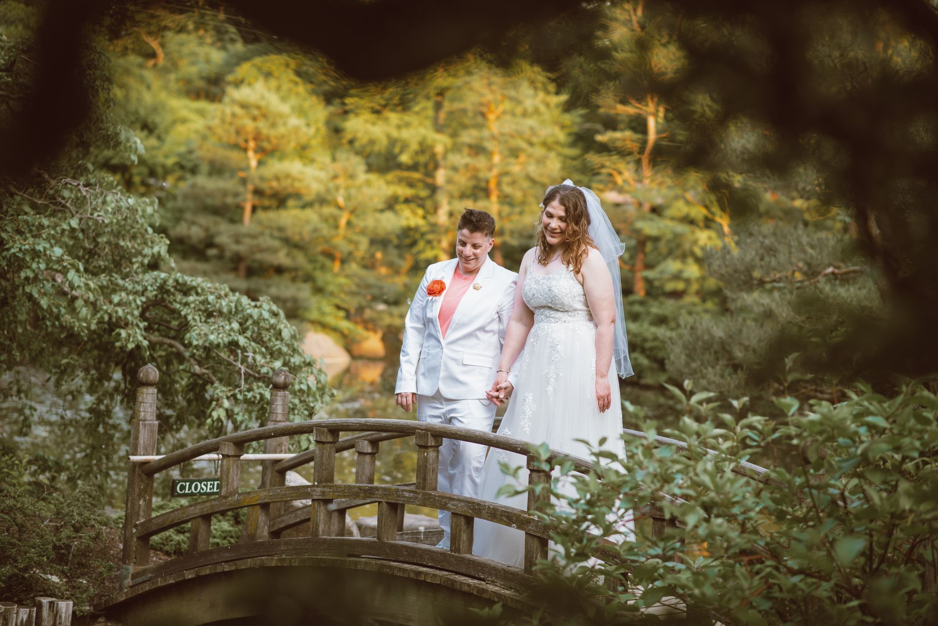 Two brides hold hands, walking on a wooden bridge, in a lush garden setting.