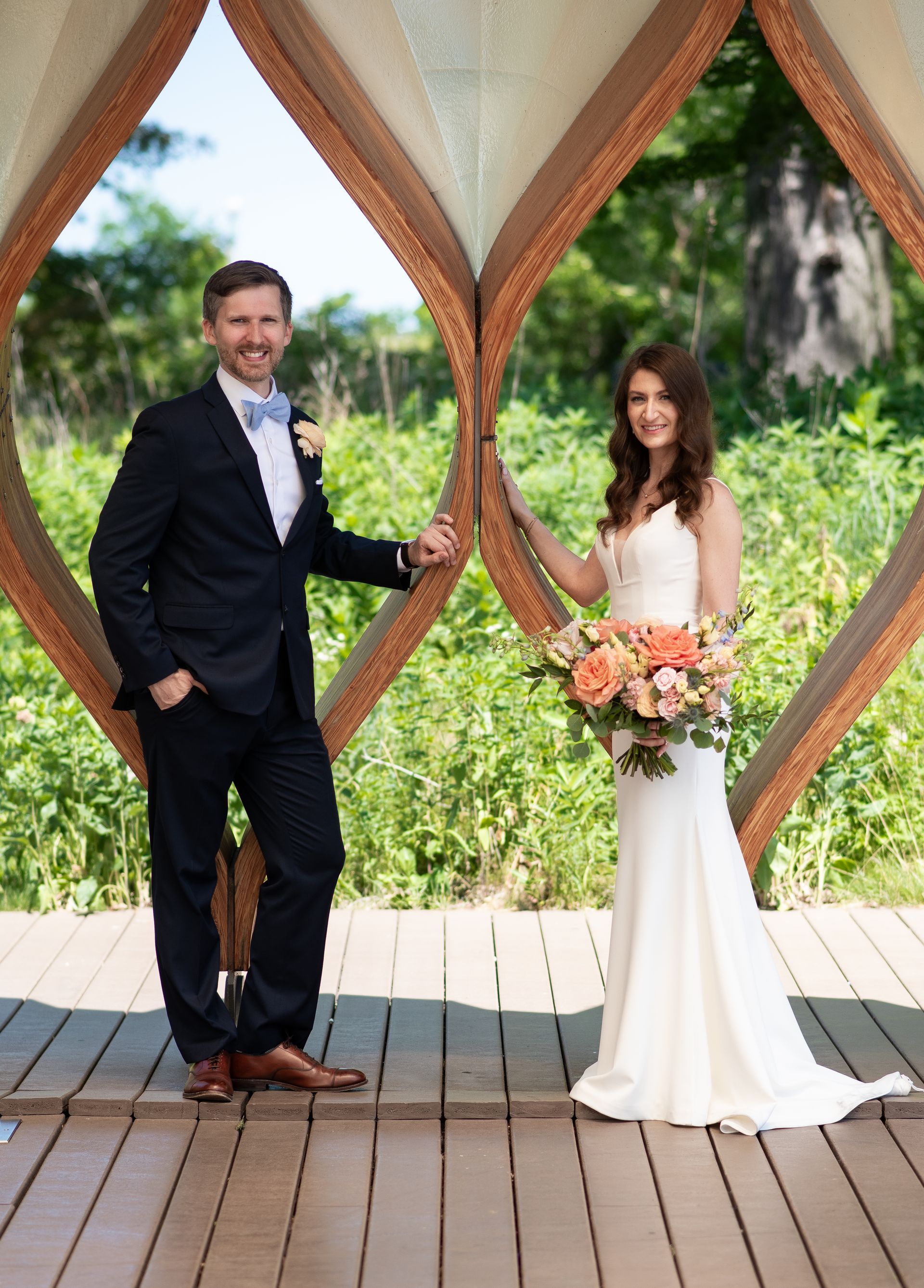 Bride and groom holding hands, smiling, standing inside a wooden structure; green foliage in the background.