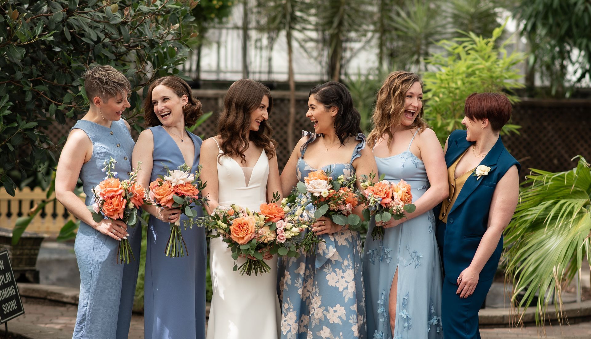 Bride with bridesmaids, smiling, holding bouquets, wearing blue dresses in a garden setting.
