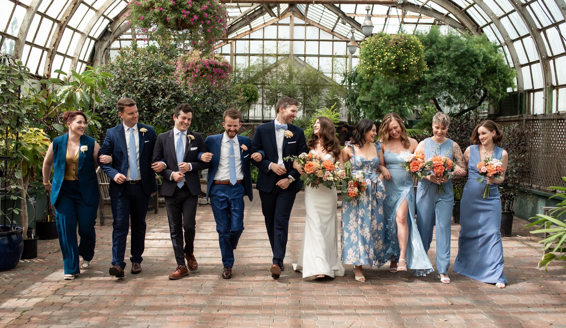 Wedding party in a greenhouse. Group walks arm-in-arm, smiling. Bride in white dress, bridesmaids in blue.