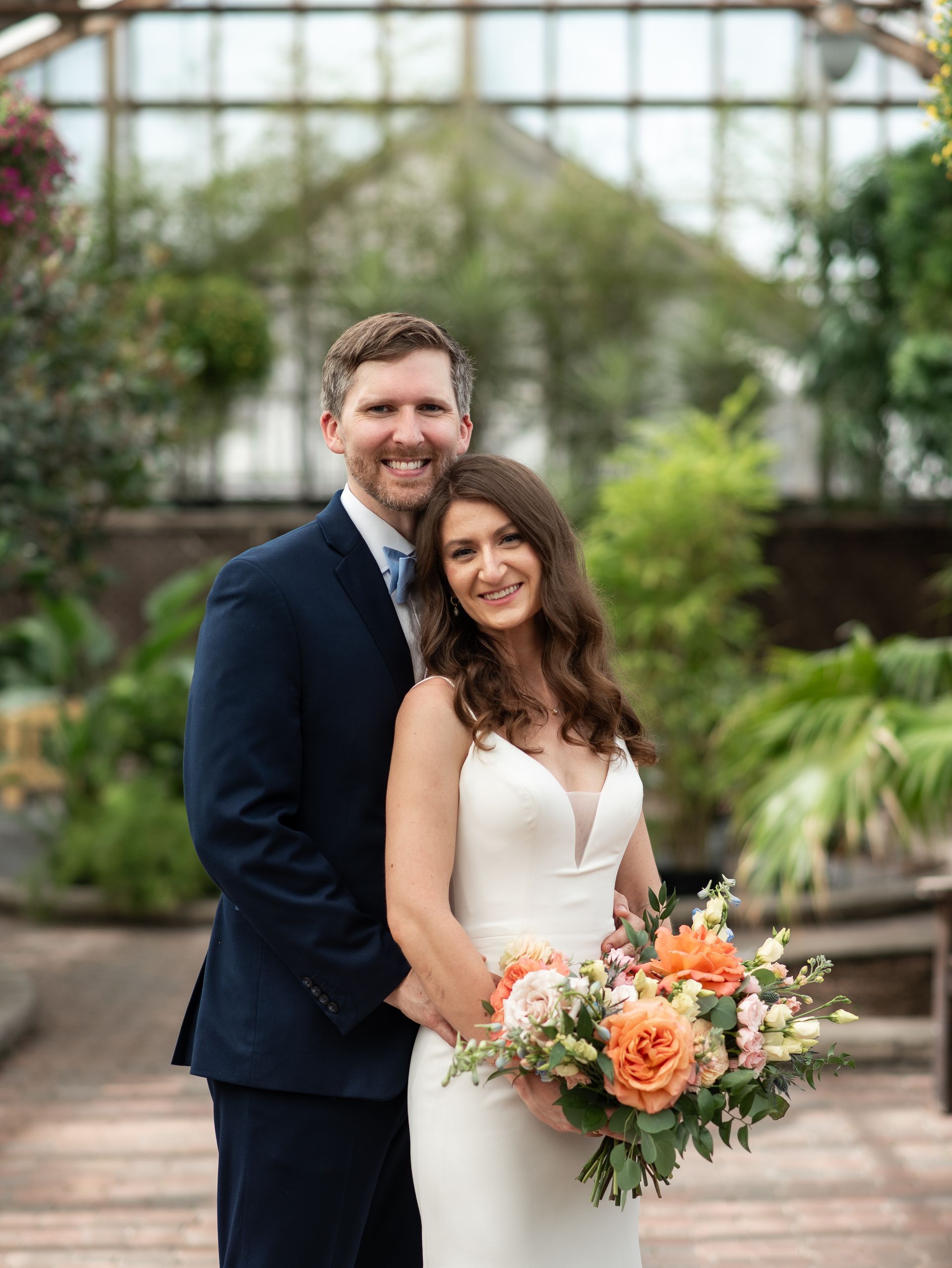 Couple smiling, embracing; woman holds bouquet; set in a greenhouse.