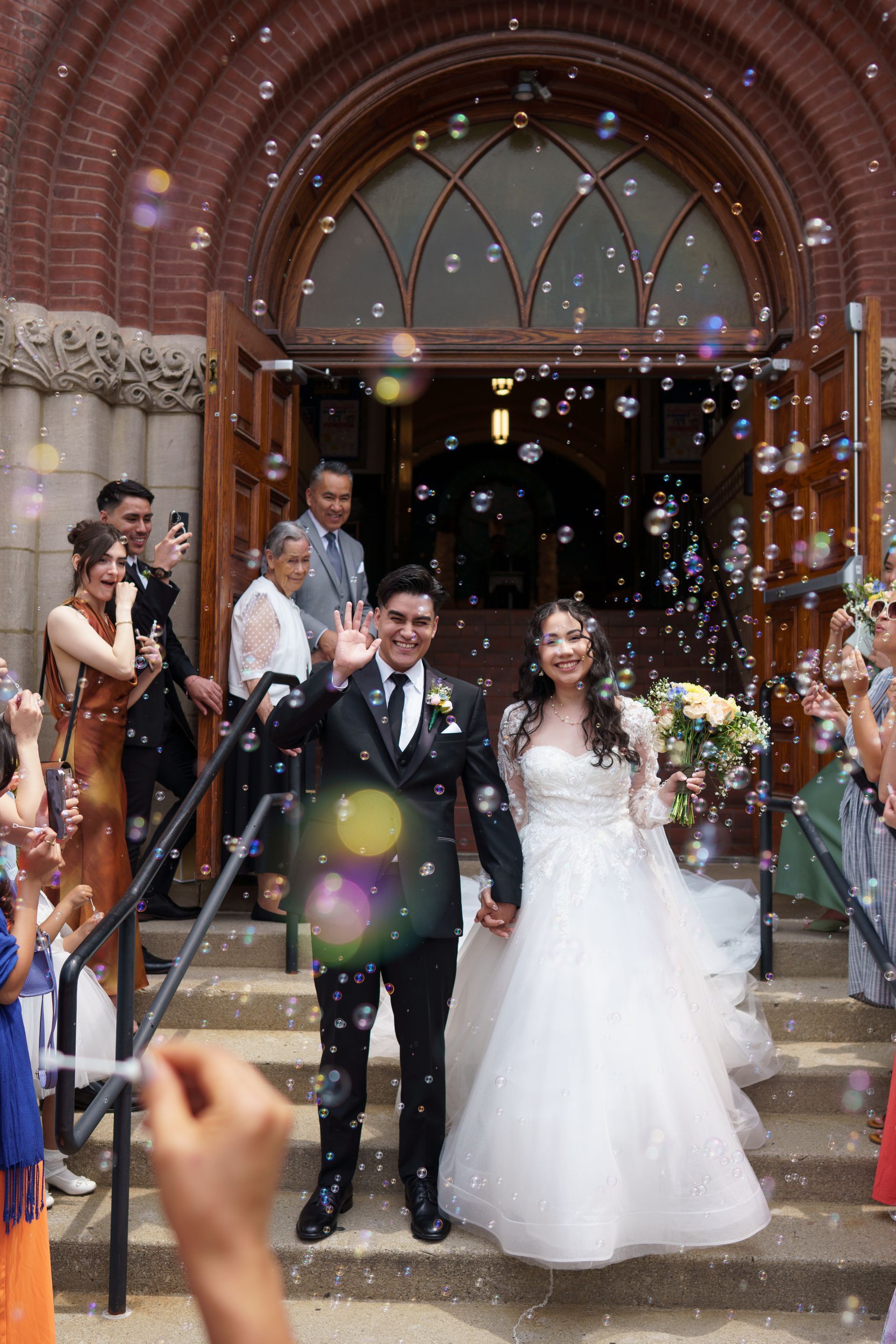 Newlyweds exit a church, waving amid bubbles. The bride wears a white gown and the groom a black suit.