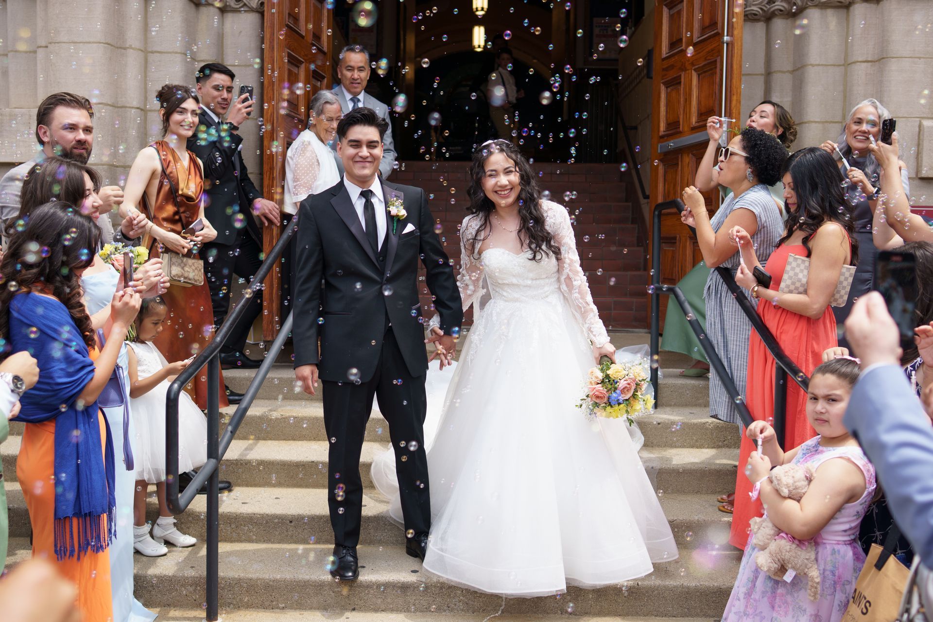 Newlyweds exiting church, smiling, surrounded by bubbles, guests cheering, and colorful bouquet.