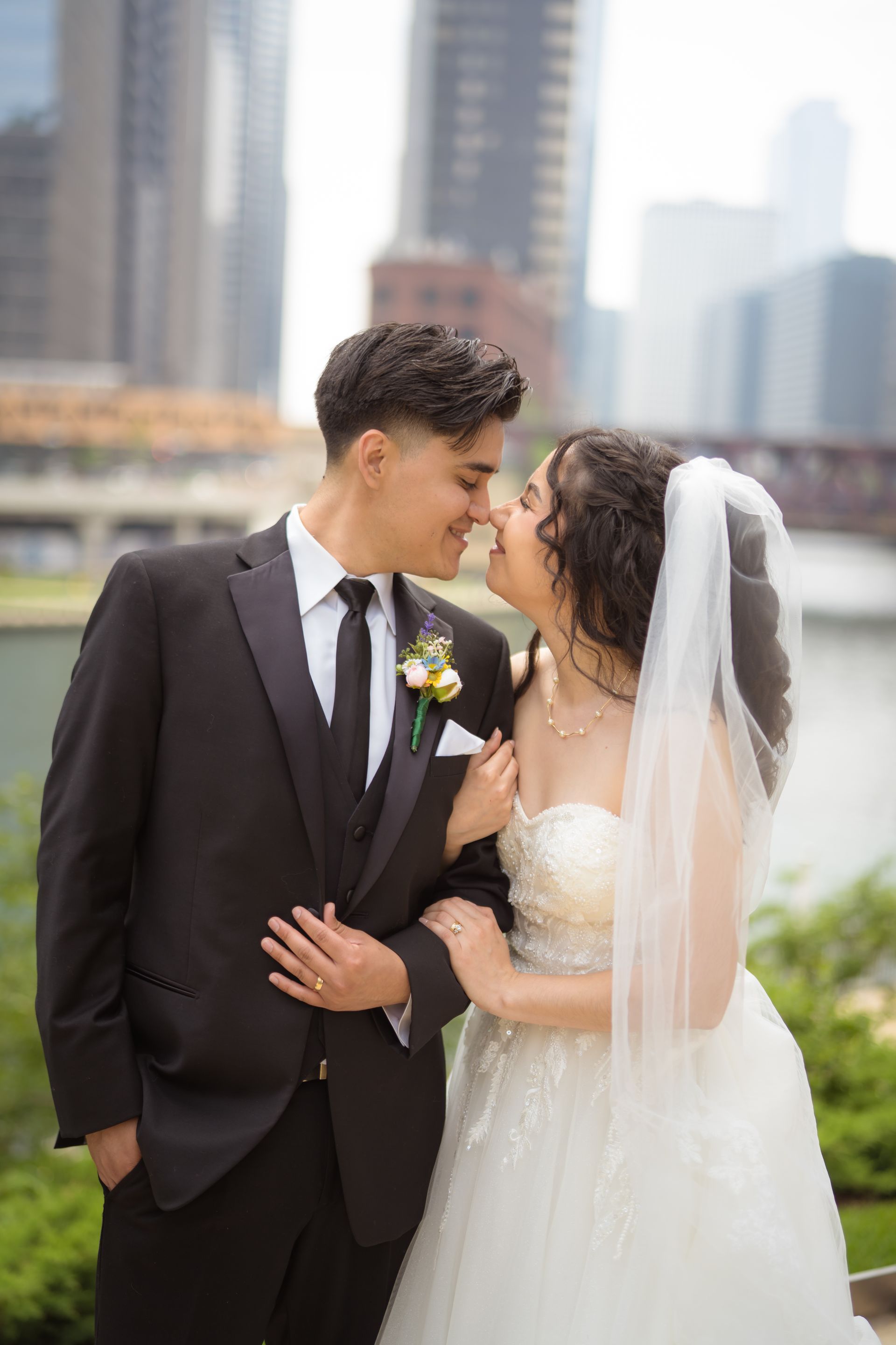 Wedding couple, smiling, about to kiss. Man in a black suit, woman in a white dress and veil, city backdrop.