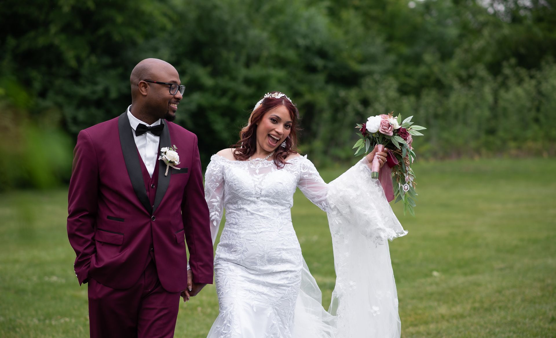 Bride and groom walk on grass, bride holding flowers, groom in burgundy suit.