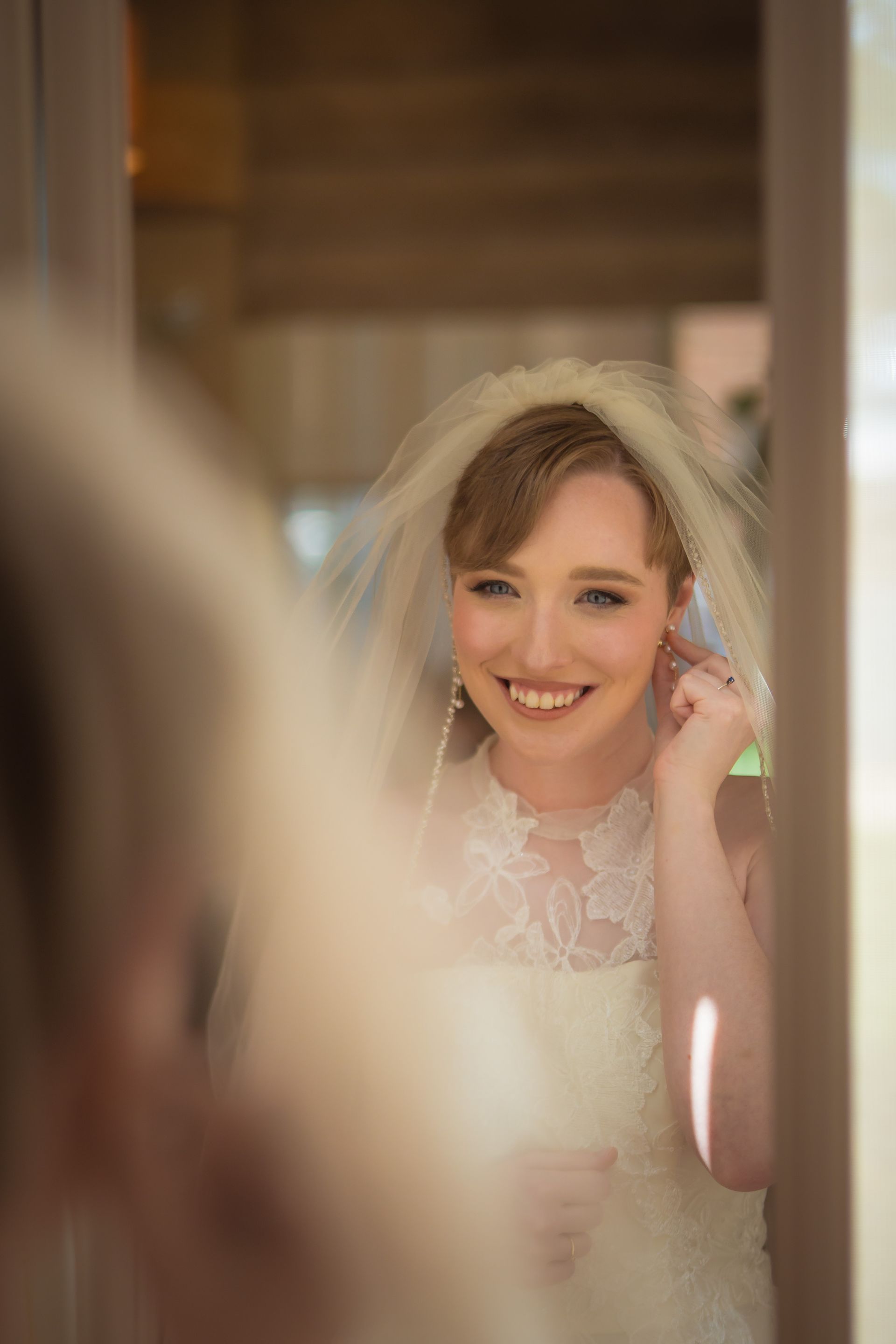 Bride wearing a veil and lace dress smiles while putting in an earring, reflected in a mirror.