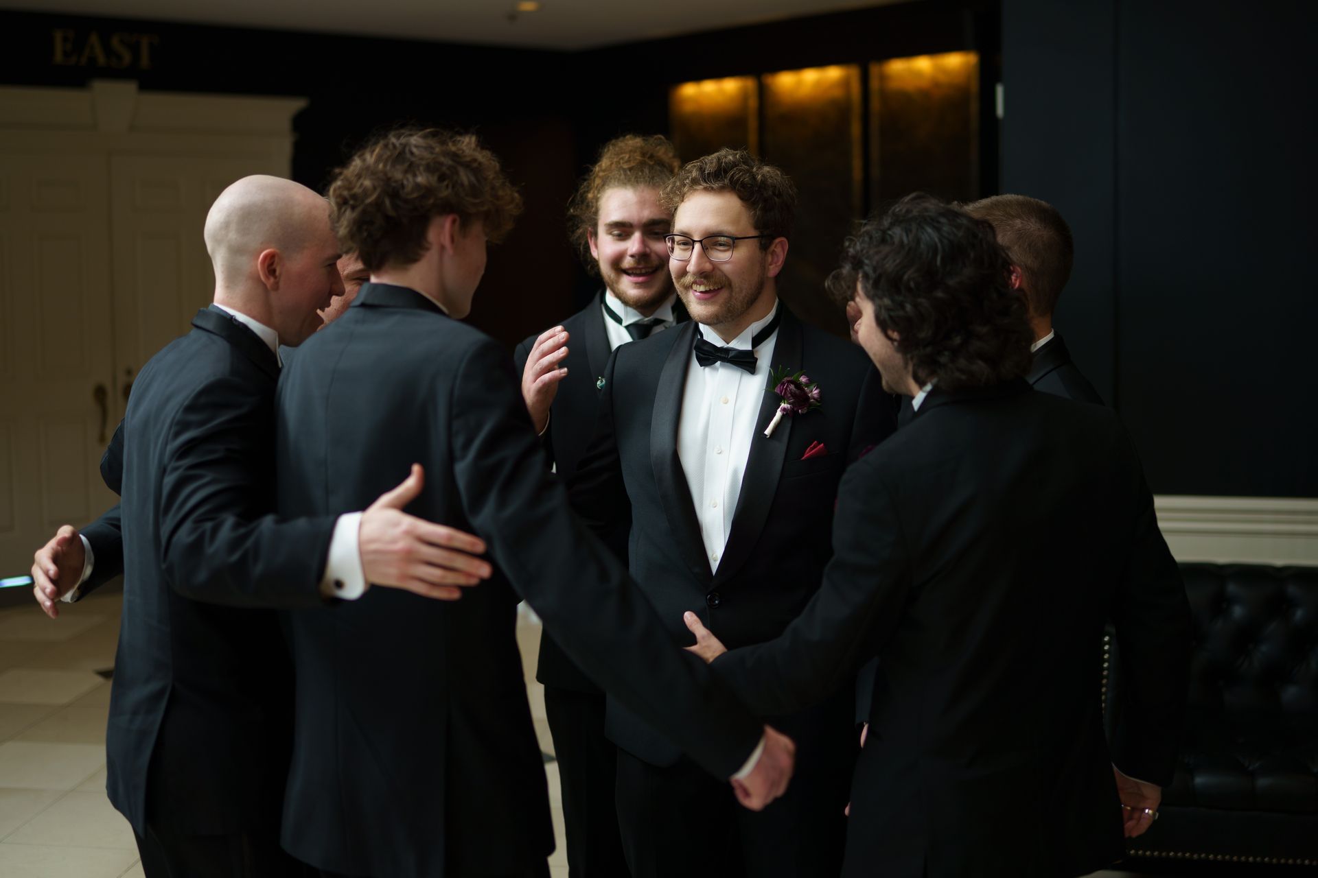 Men in tuxedos embrace, likely congratulating each other, in a formal interior setting.