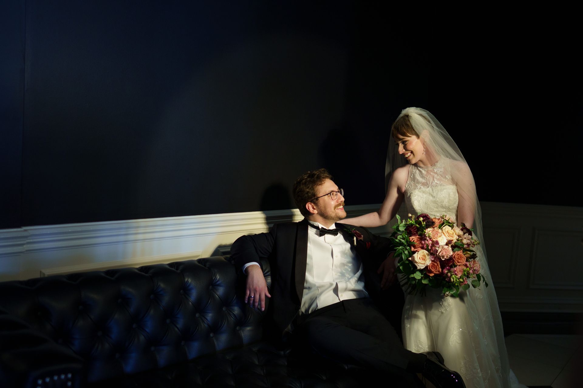 Bride in white gown touches groom's shoulder while he sits on a black couch, both smiling. Dark background.