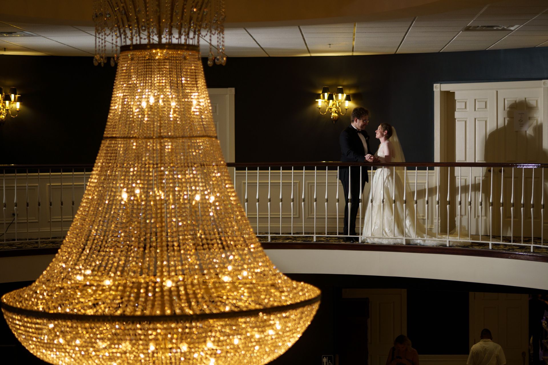 Bride and groom on a balcony, illuminated by a large chandelier in a ballroom setting.