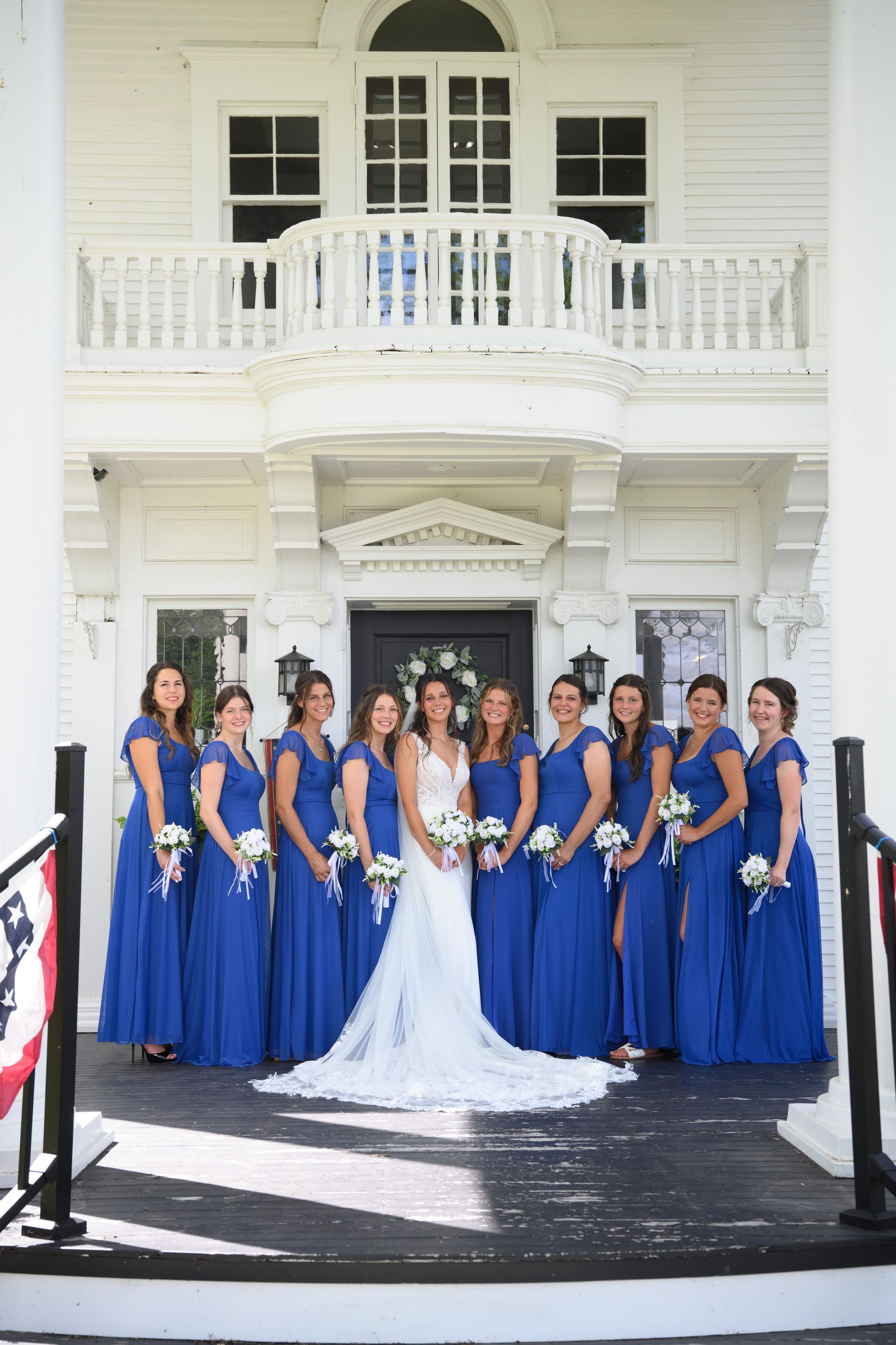 Bride and bridesmaids in blue dresses pose on a porch in front of a white building.