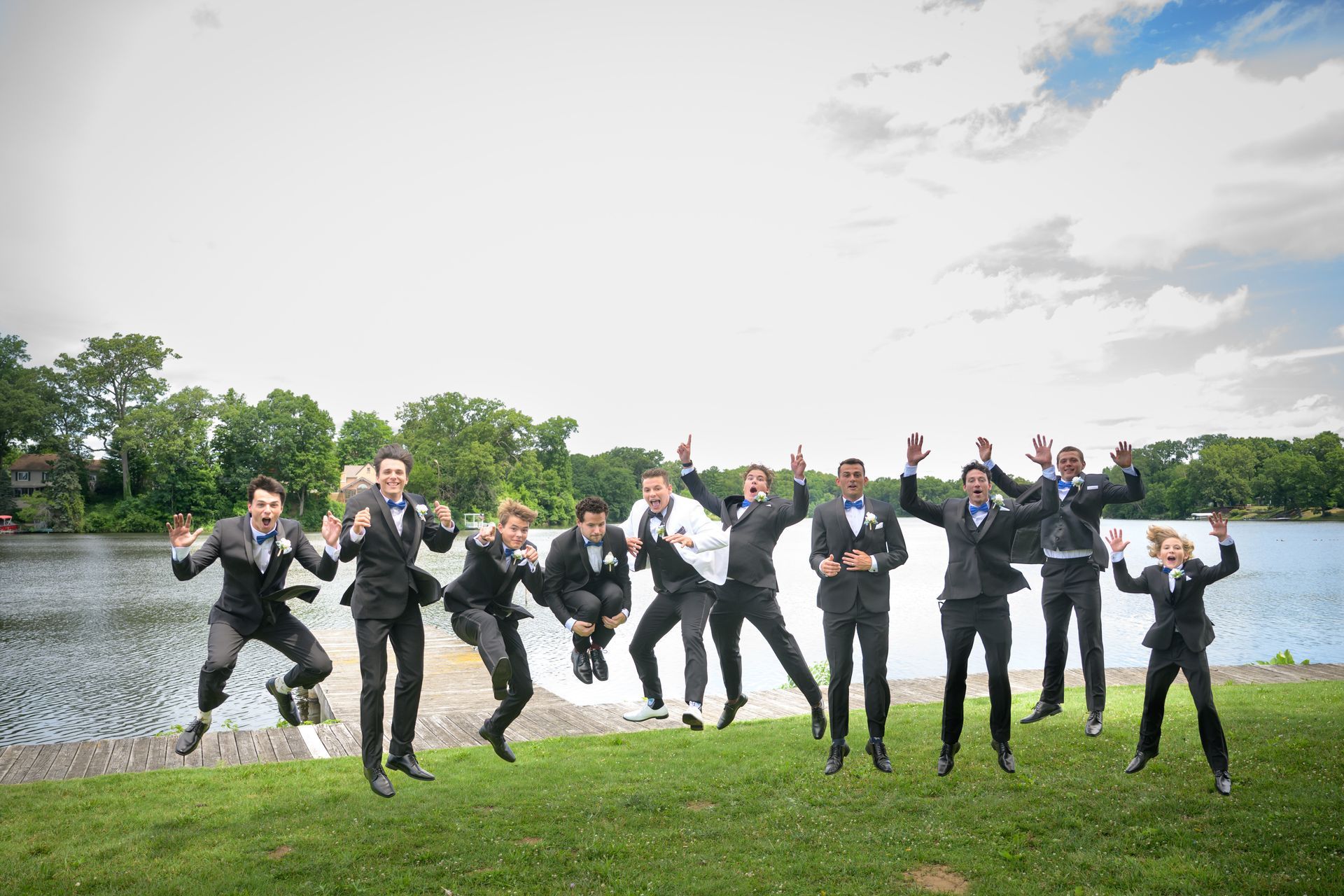Men in black suits jump in the air on a grassy area, with a lake and trees in the background.