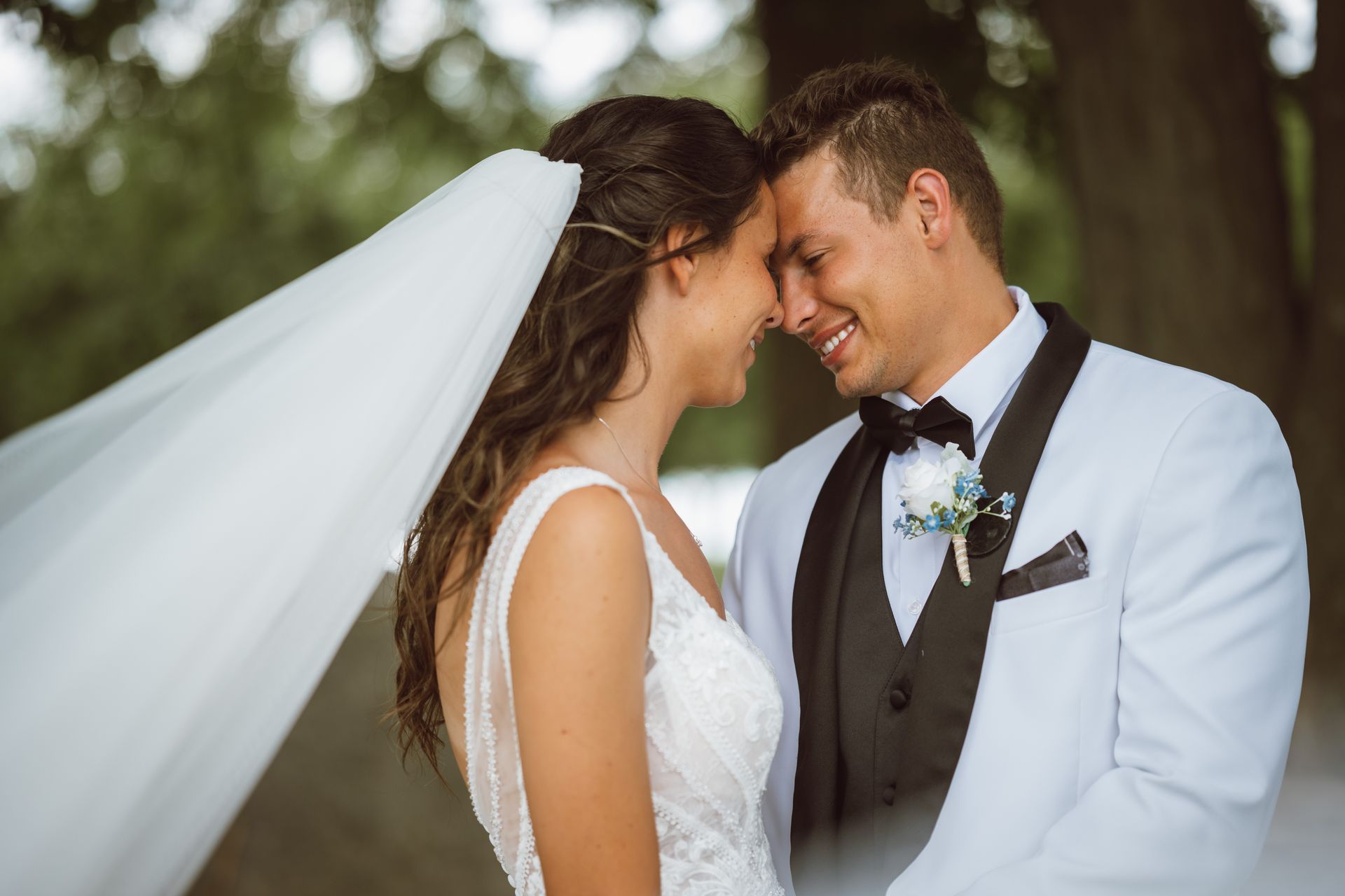 Bride and groom smiling, touching foreheads. Bride in white dress and veil, groom in white suit and black bow tie, outdoors.